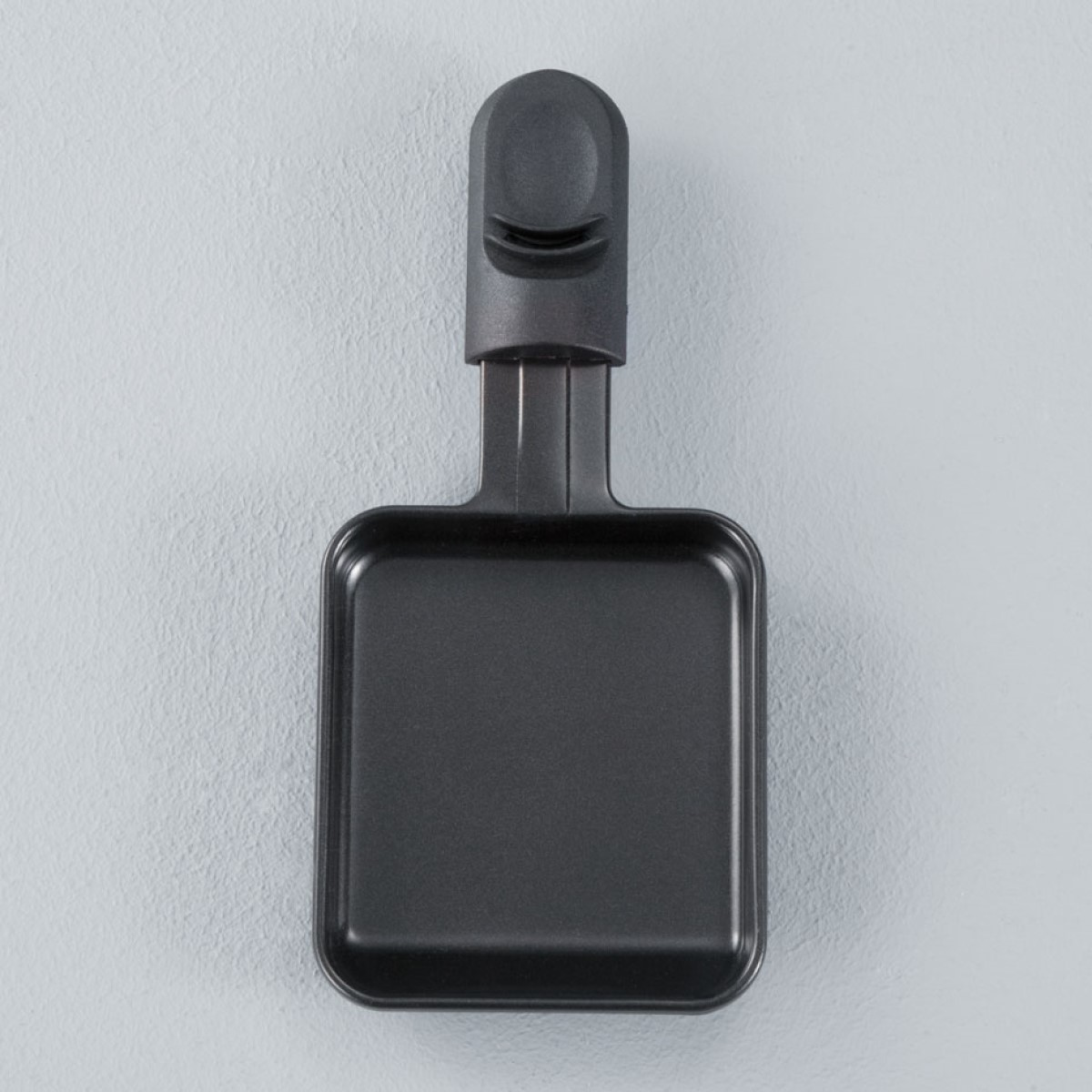 Black raclette grill pan with a handle, viewed from above. Set against a light gray surface.
