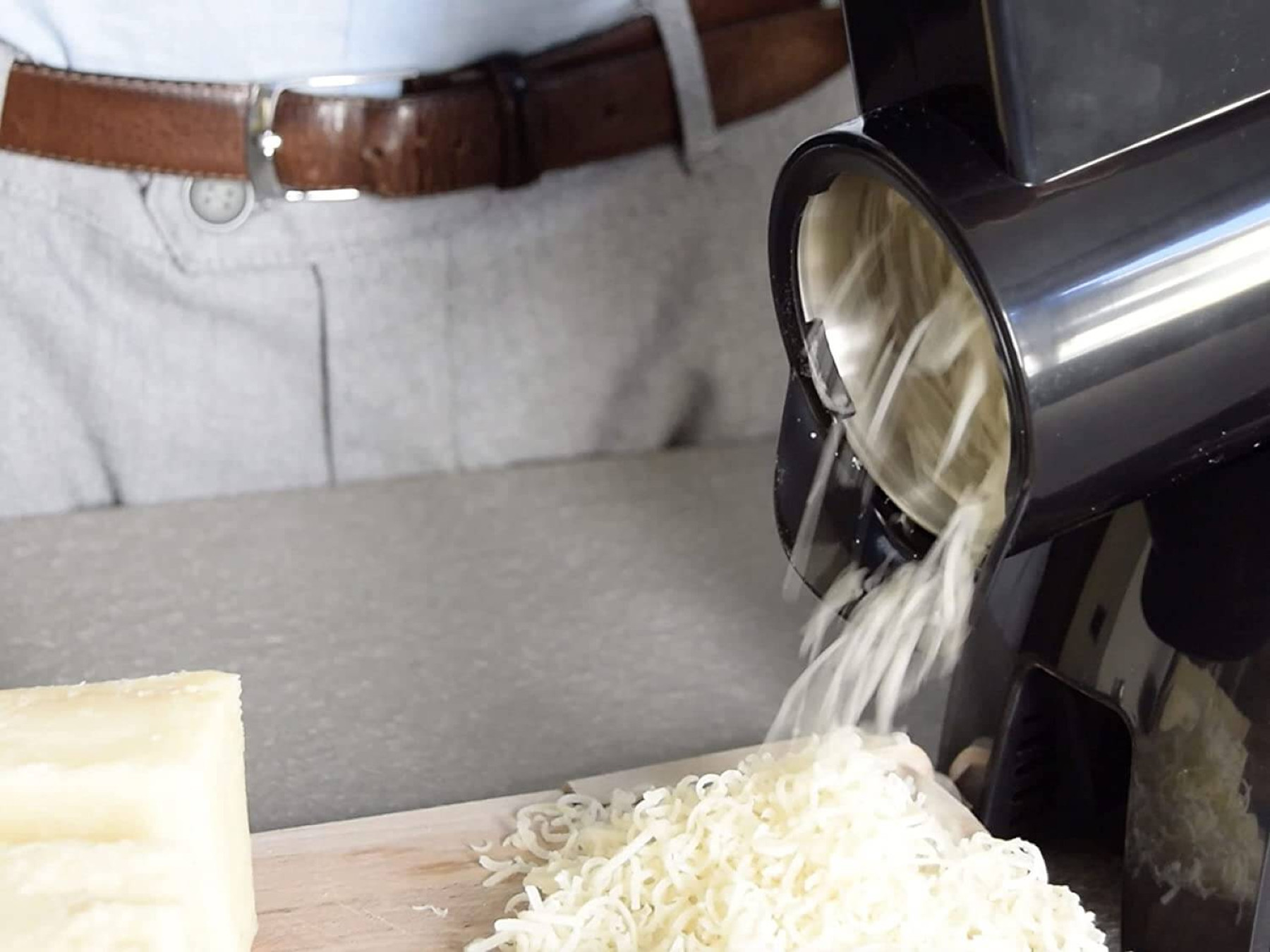 Eine Person reibt Käse mit einer Maschine. Käse fällt auf eine Oberfläche.