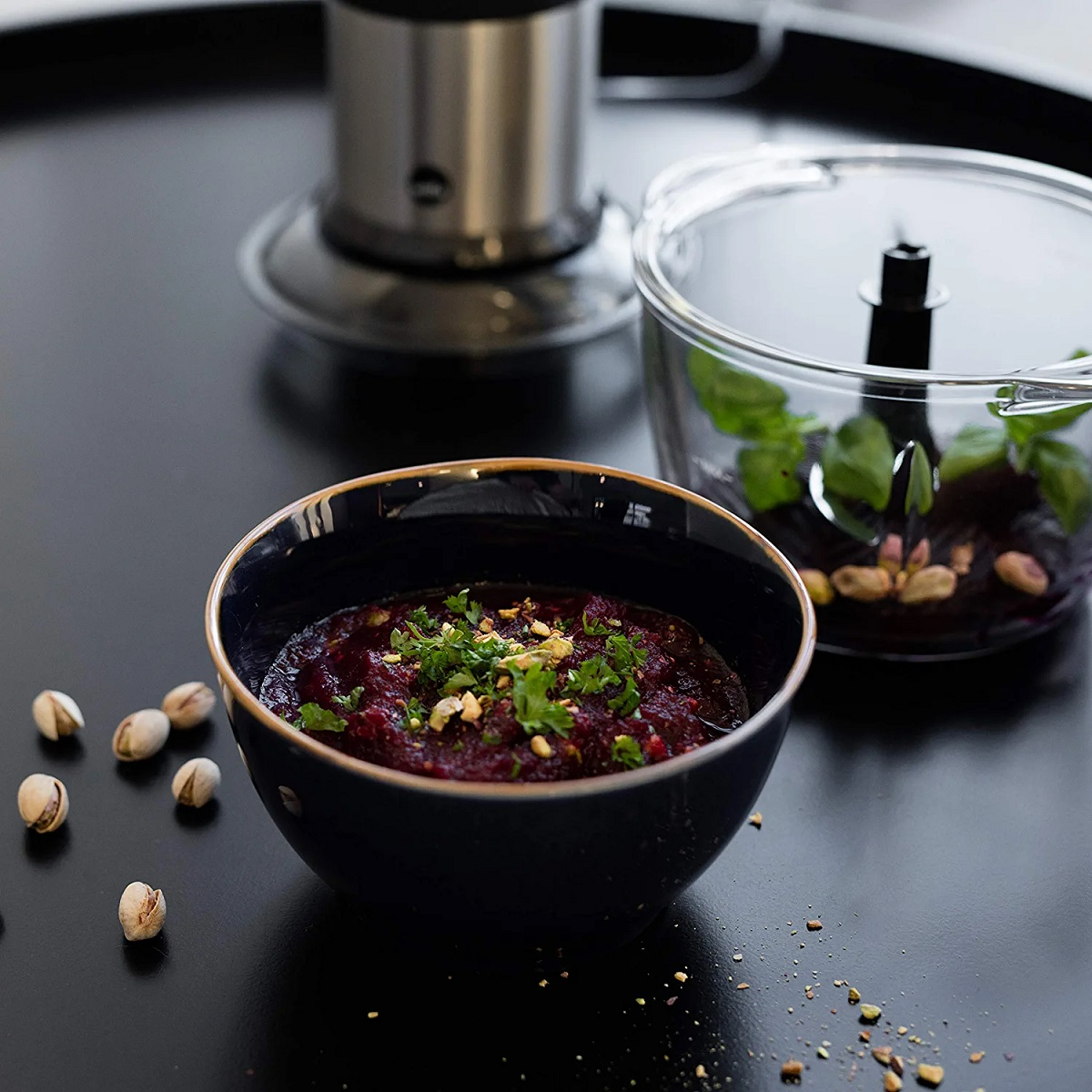 Nahaufnahme einer schwarzen Schüssel mit rotem Dip, Pistazien und Kräutern auf schwarzem Untergrund. Klare Schüssel sichtbar.
