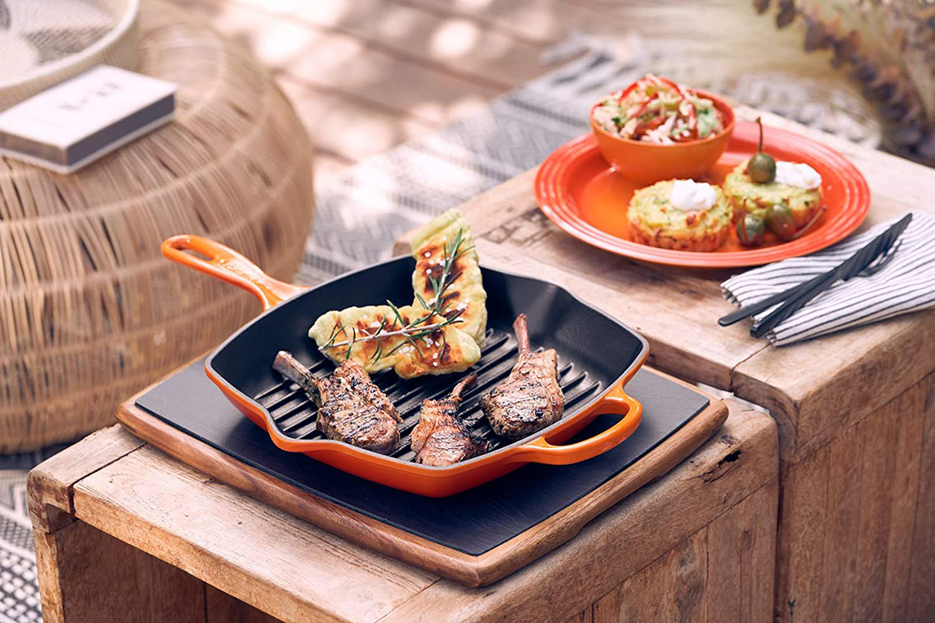 Gegrillte Lammkoteletts auf einer schwarzen Pfanne auf einem Holztisch, mit Beilagen und einem gewebten Hocker.