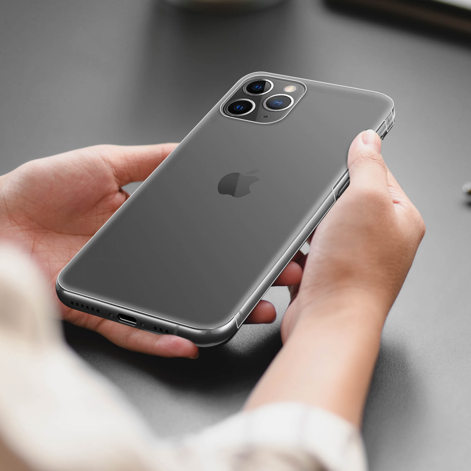 Una persona tiene in mano un iPhone grigio scuro con una custodia trasparente. Il logo Apple è visibile. Lo sfondo è una superficie grigia.
