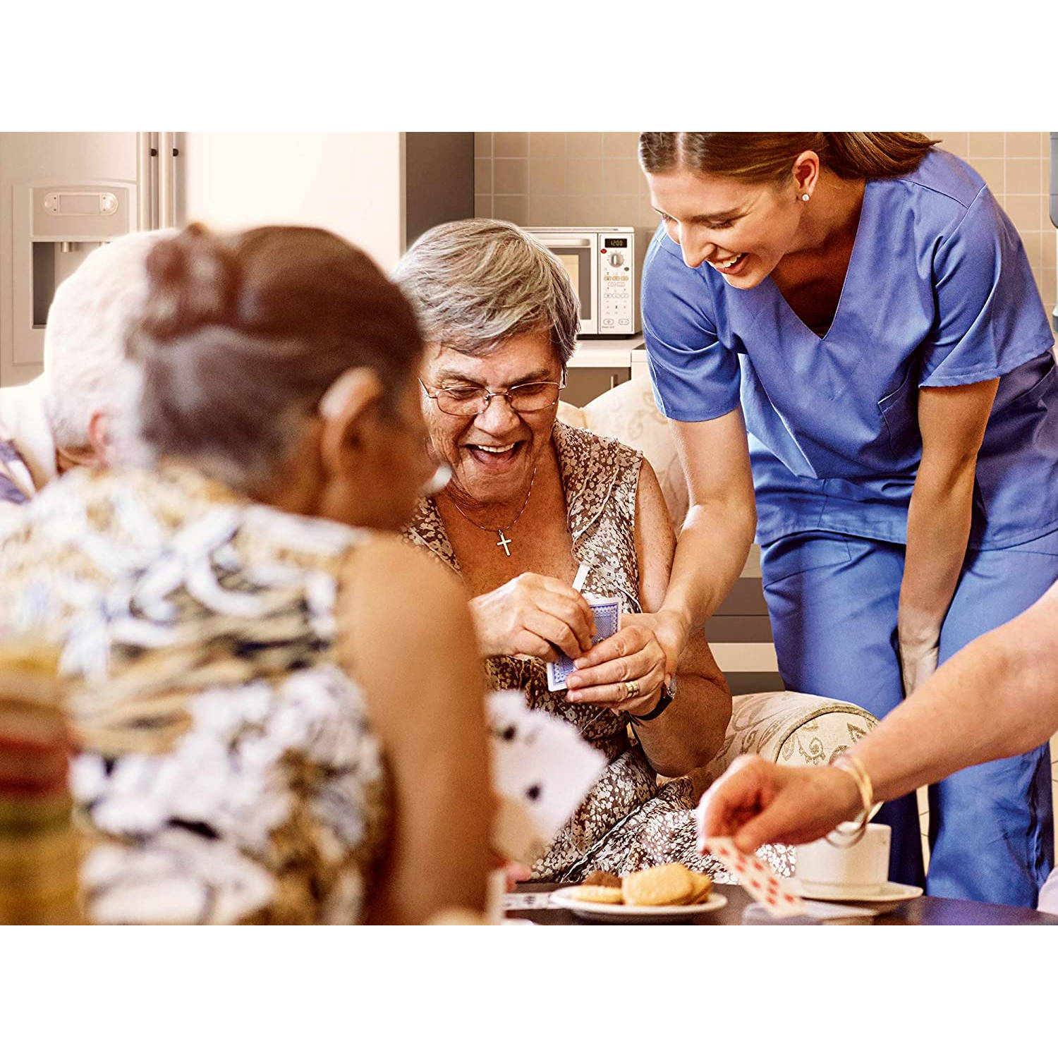 Un'infermiera che aiuta gli anziani a giocare a carte. Persone sorridenti a un tavolo con carte, biscotti e microonde.