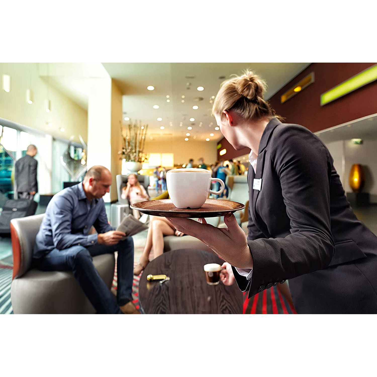 Una cameriera che serve caffè nella hall di un hotel. Persone sullo sfondo.