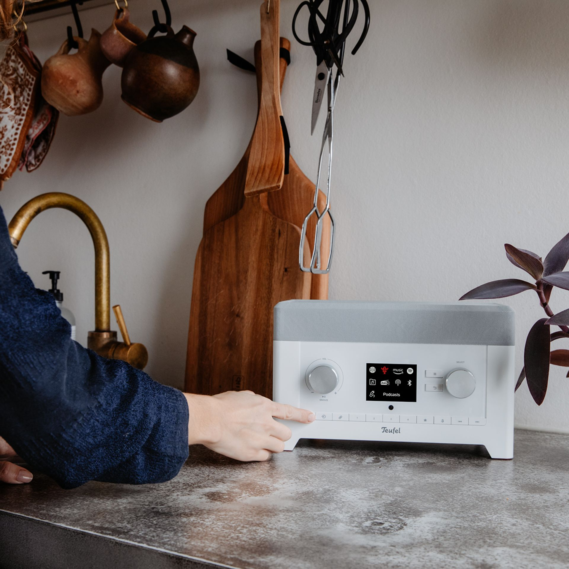 Een witte en grijze Teufel radio op een aanrecht. Een hand drukt op een knop. Keukenomgeving.