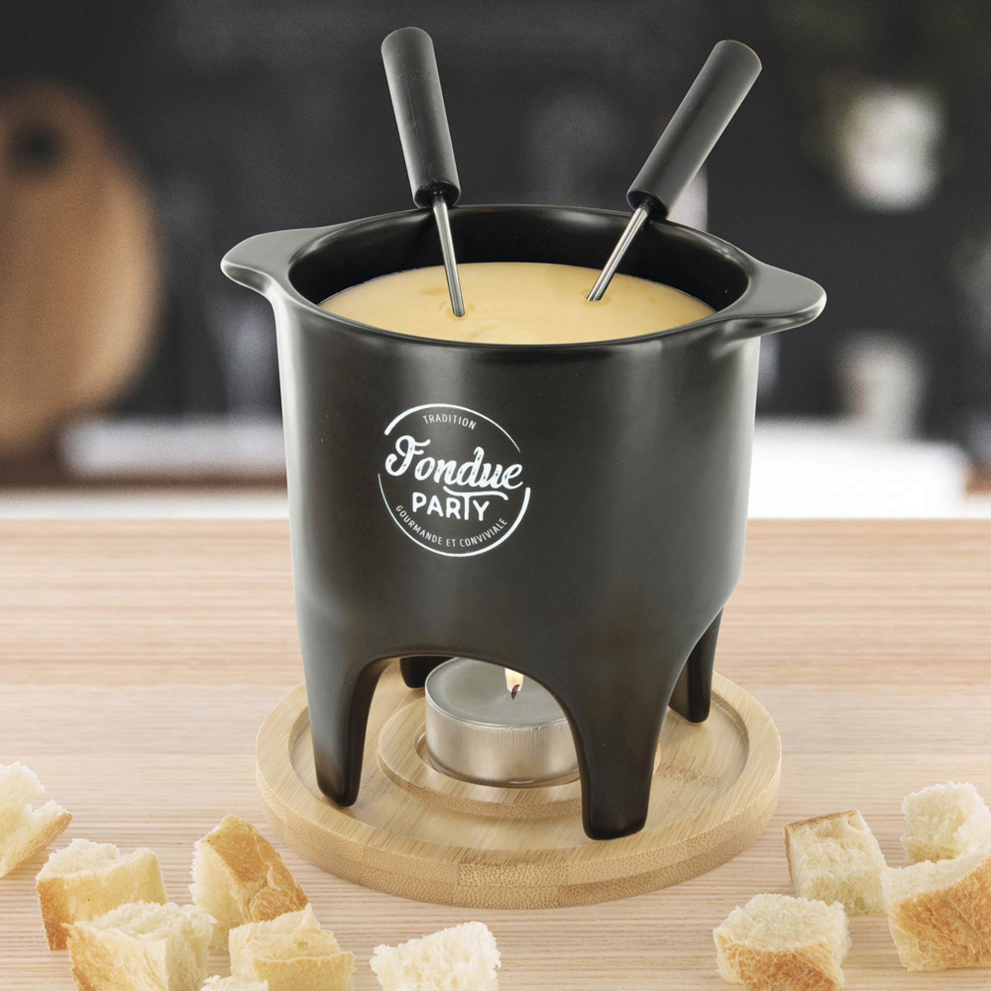Pentola per fonduta nera con due forchette, una candela e cubetti di pane su base di legno.