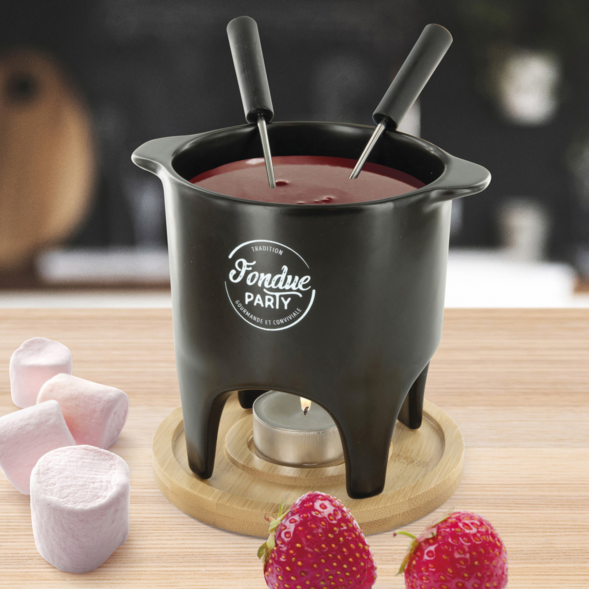 Pentola per fonduta nera con liquido rosso, due forchette, marshmallow, fragole e una candela.
