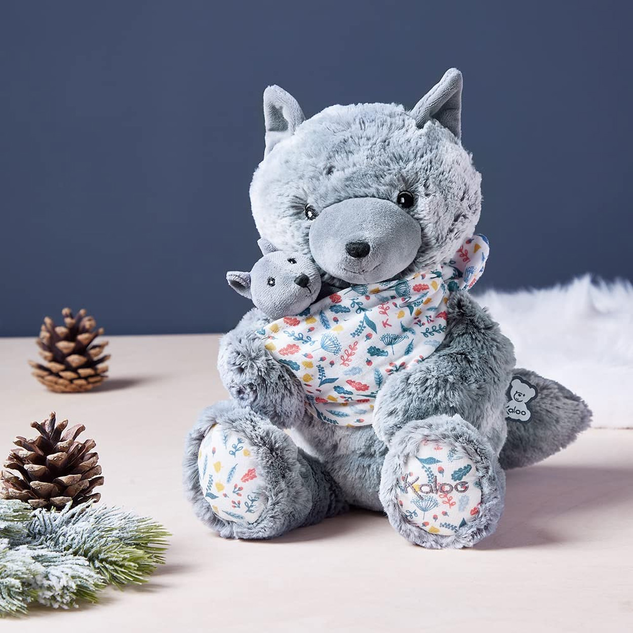 Une peluche de loup gris tenant un jouet de loup plus petit. Ils sont sur une surface claire.