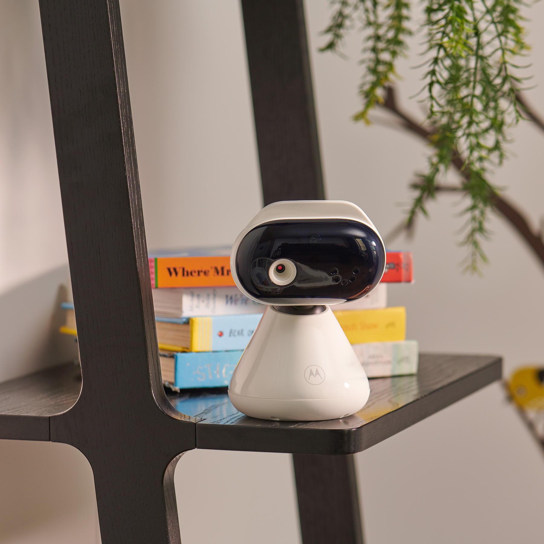 Weißer Babyphone auf einem Regal mit Büchern und einer Pflanze, schwarze Leiter im Hintergrund.