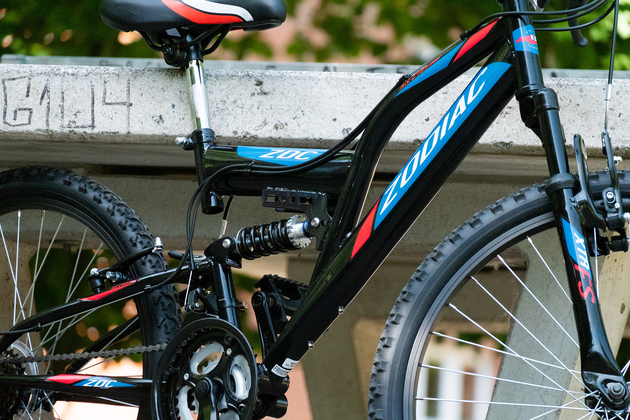 Schwarzes und blaues Zodiac Mountainbike mit gefedertem Rahmen, auf Betonfläche, grüne Bäume im Hintergrund.