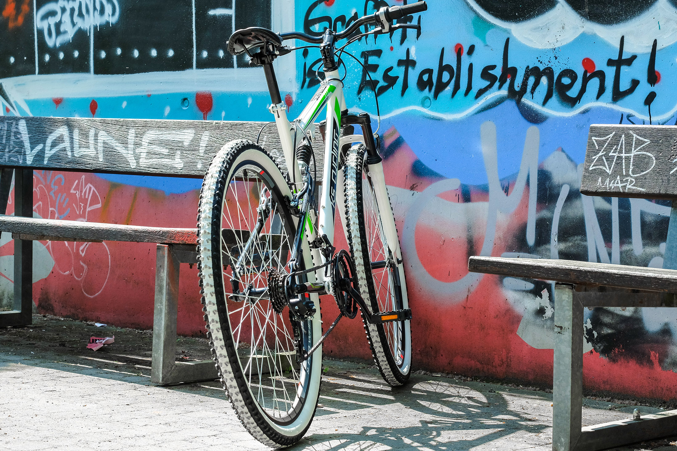 Weiß-grünes Mountainbike lehnt an einer Wand mit Graffiti. Die Szene spielt im Freien an einem sonnigen Tag.