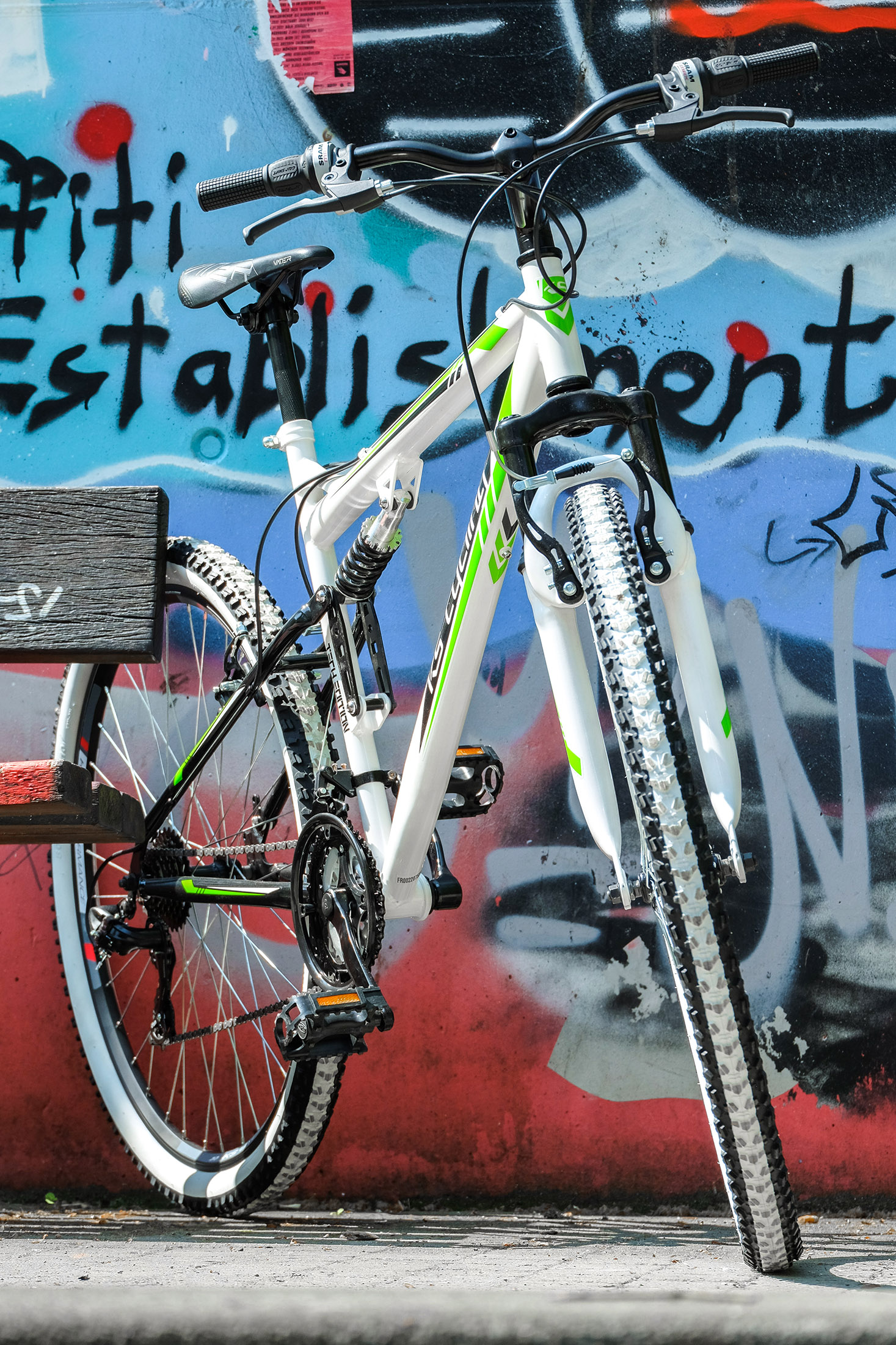 Ein weißes und grünes Fahrrad steht vor einer Graffiti-Wand. Das Fahrrad steht auf einer Straße.