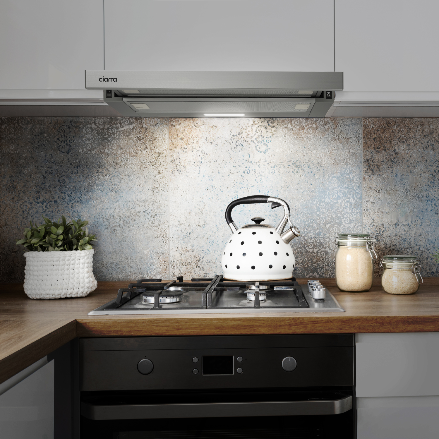 Kitchen scene: Range hood, gas stove, kettle, jars, plant. Background: patterned backsplash.