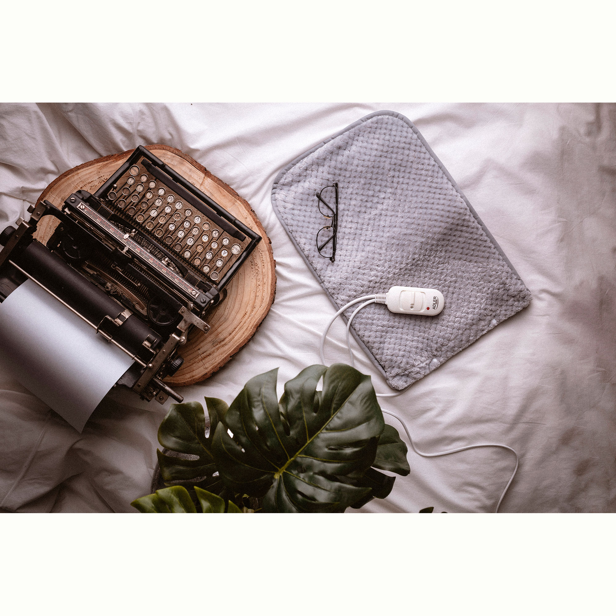 Una máquina de escribir, una almohadilla térmica y gafas descansan sobre una cama. Una planta debajo.