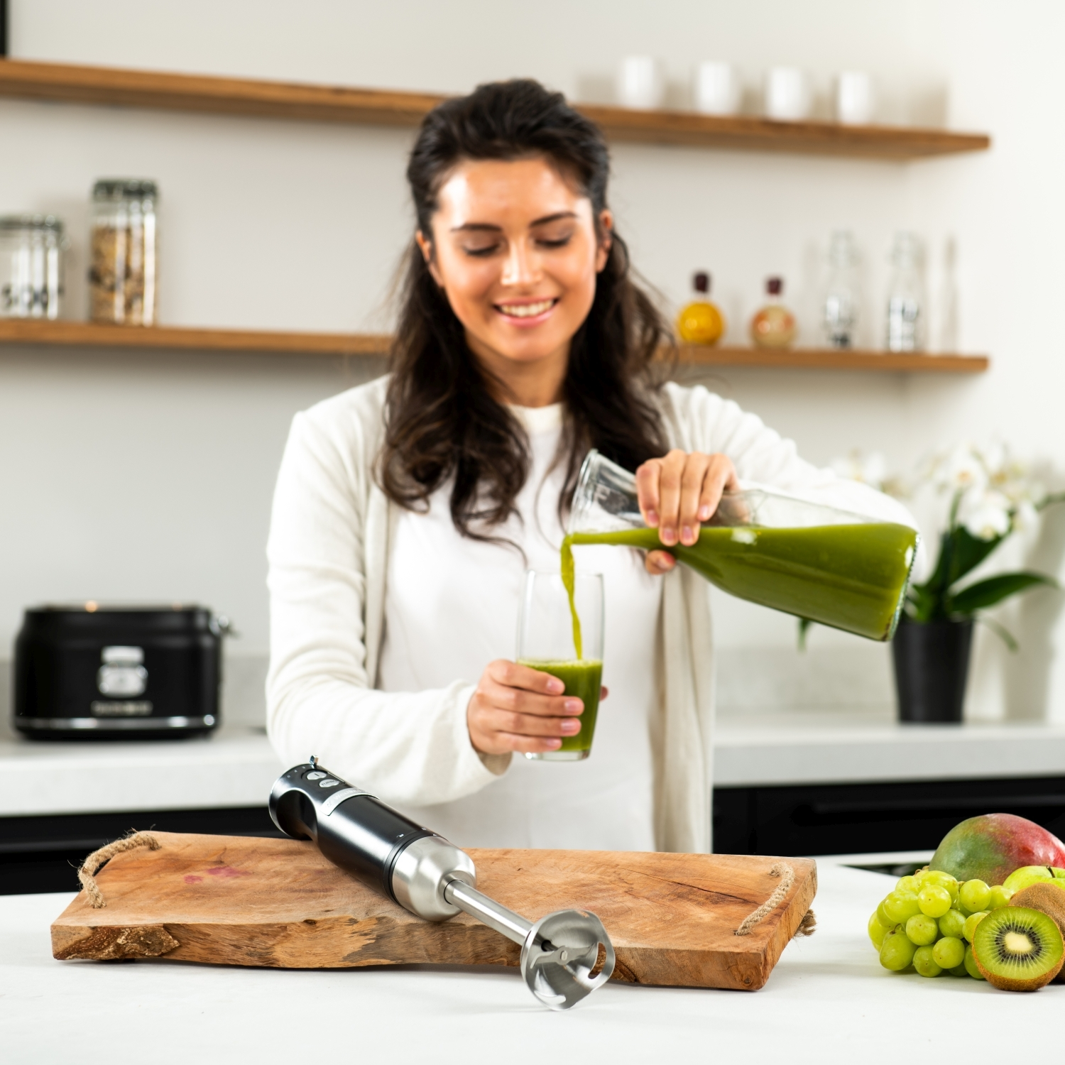 Frau macht Saft. Sie gießt Saft in ein Glas. Auf dem Tisch liegen Früchte und ein Mixer.