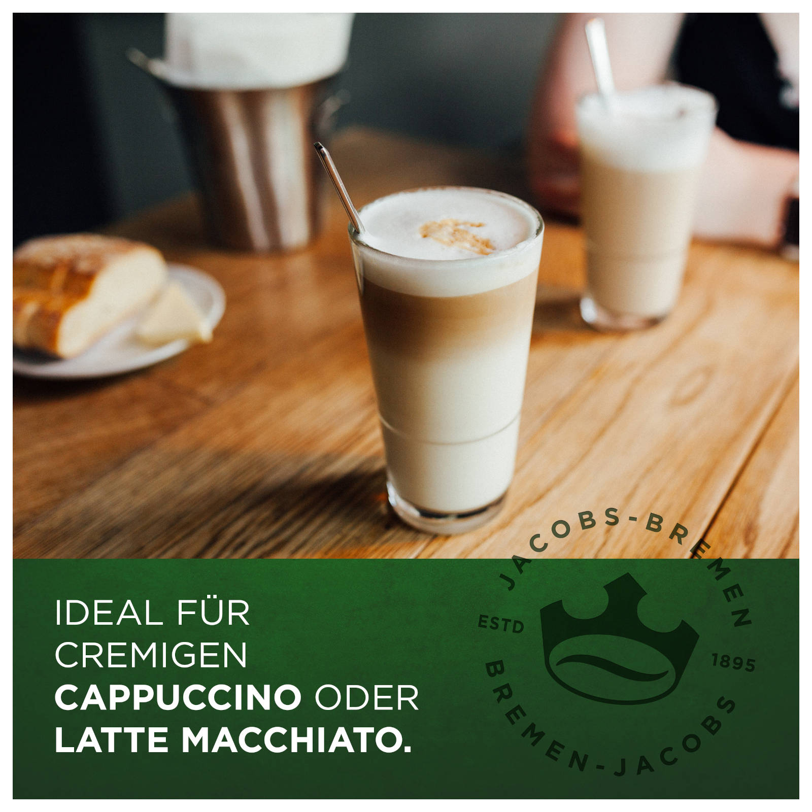 Ein Cappuccino in einem hohen Glas mit einem Löffel. Im Hintergrund ist ein weiterer Latte.