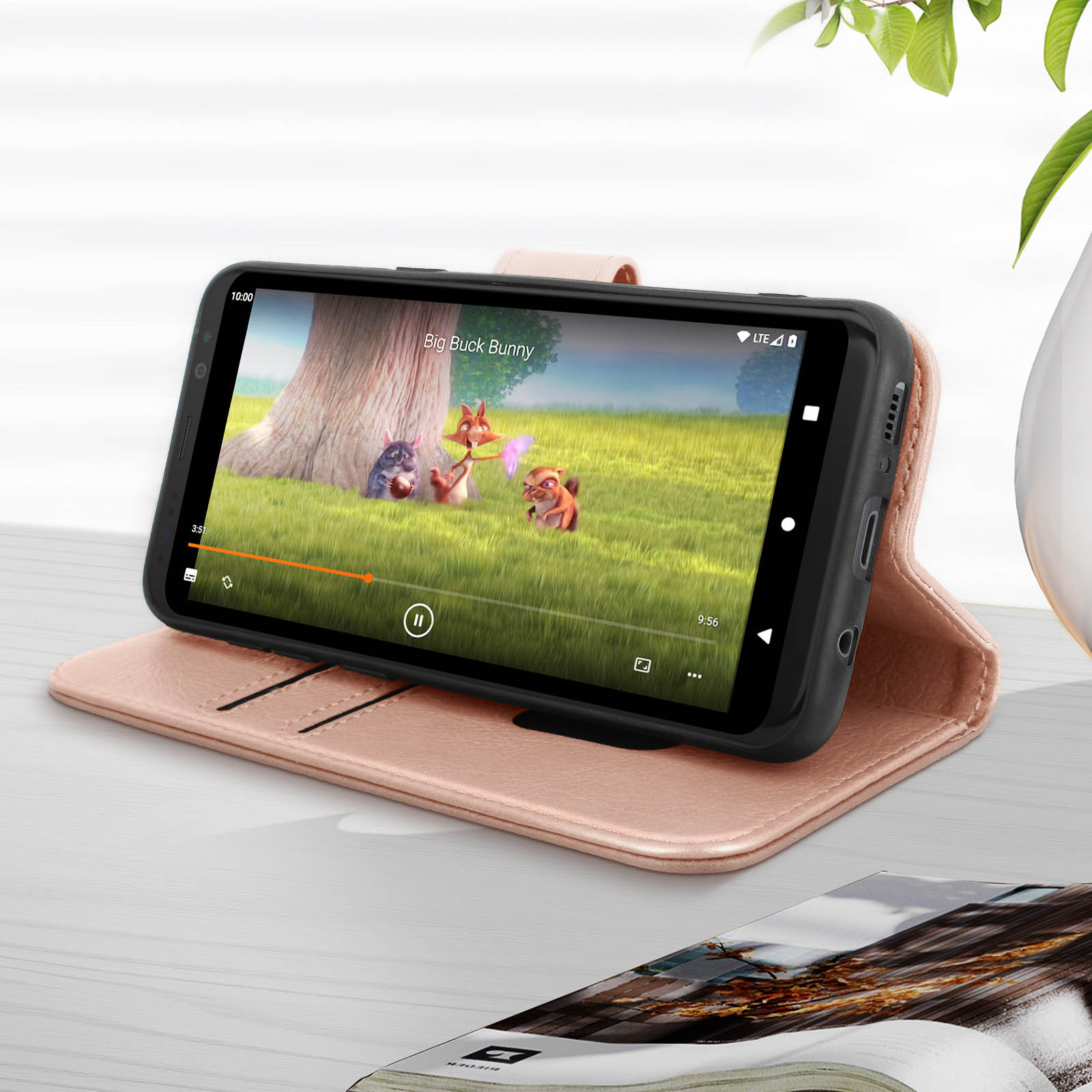 Le téléphone affiche une scène de dessin animé. Il est sur un étui rose, sur une table en bois. Un magazine est au premier plan.