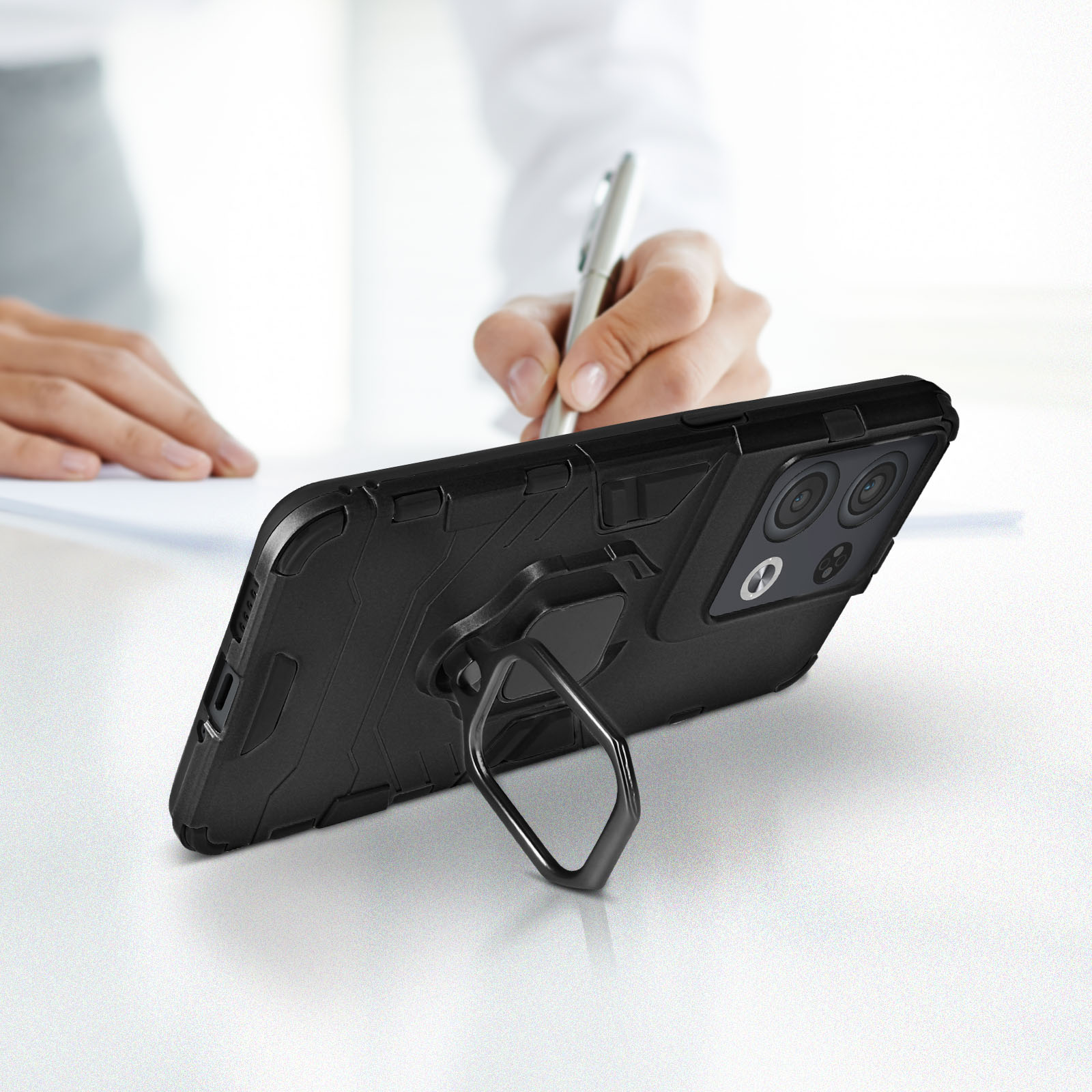 Coque de téléphone noire avec anneau sur table blanche, personne écrivant en arrière-plan.