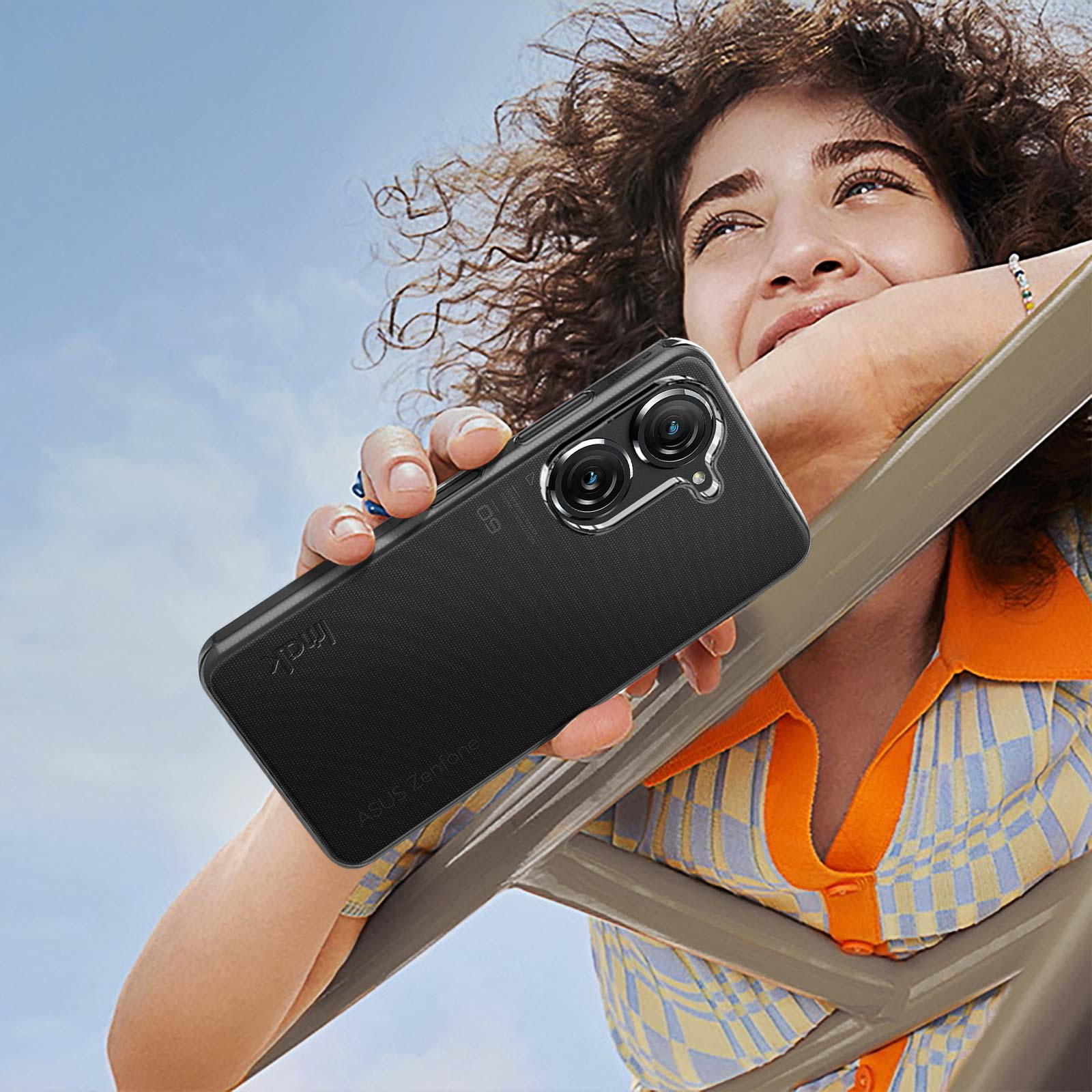 Donna che tiene telefono con custodia nera, volto rivolto verso l'alto, cielo soleggiato.