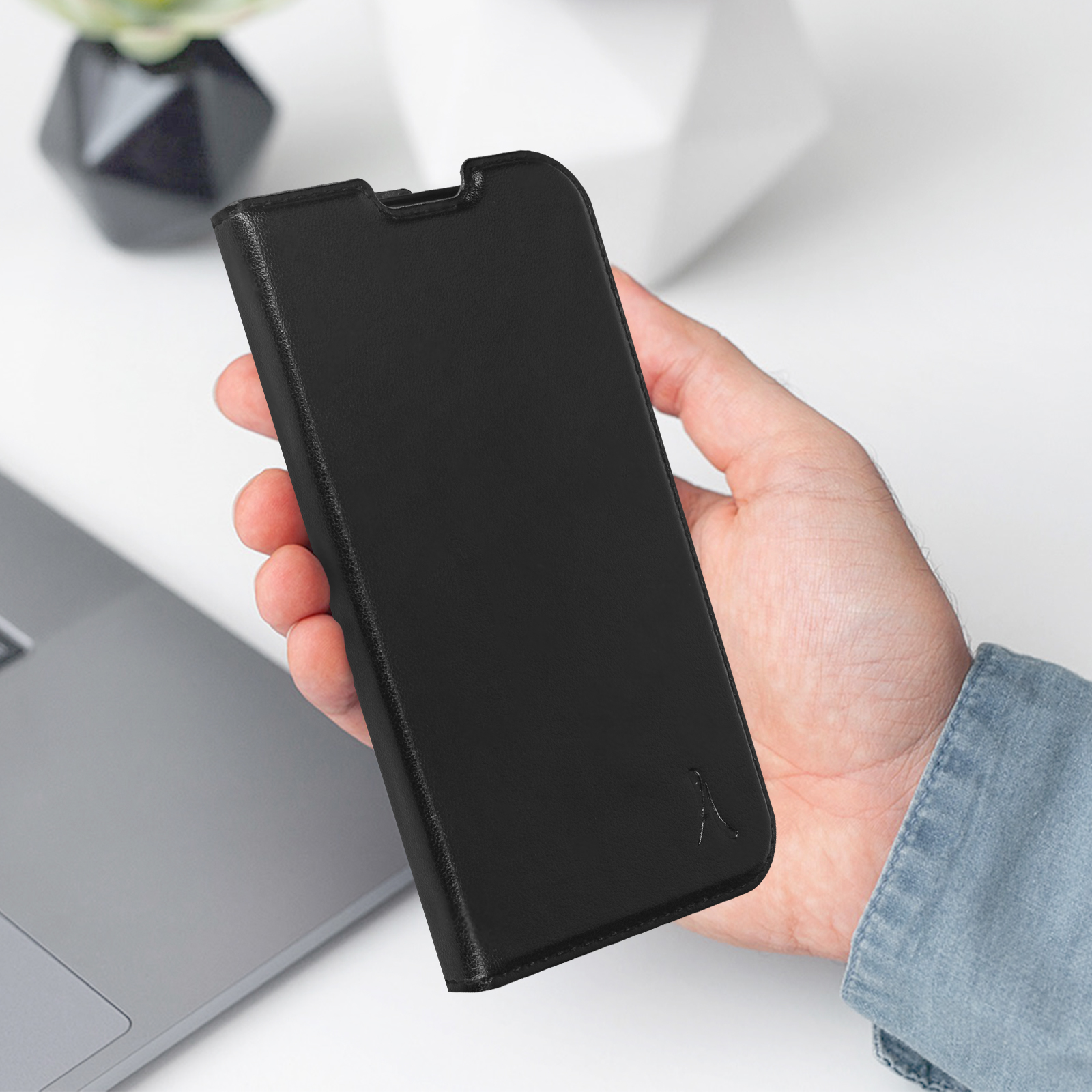 Una mano che tiene una custodia per telefono in pelle nera. Un laptop e decorazioni sullo sfondo.