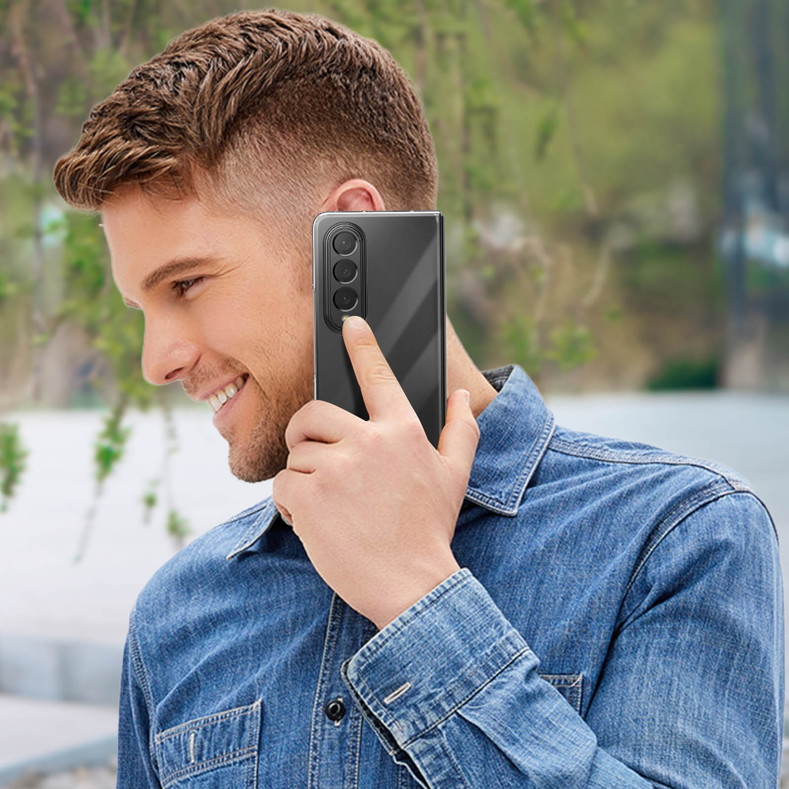 Un homme souriant tenant un téléphone noir à son oreille. Il porte une chemise en jean. Arrière-plan flou.