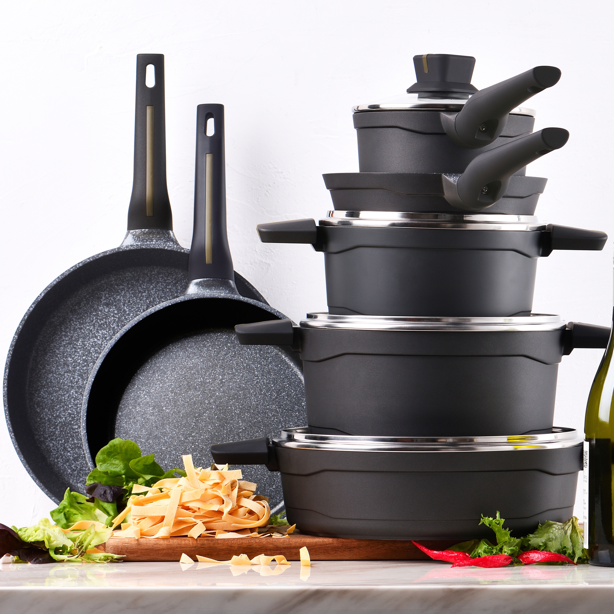 Juego de utensilios de cocina negros con sartén, ollas apiladas y tabla de madera con verduras.
