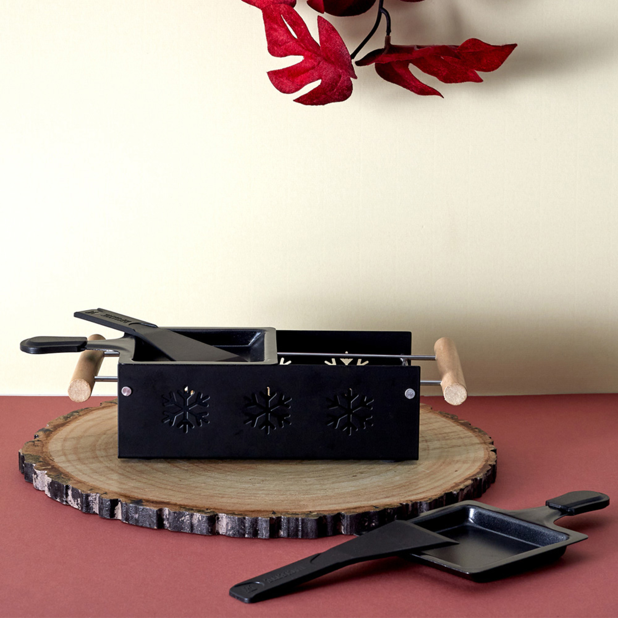 Griglia per raclette nera su una fetta di legno, padelle per raclette, foglie rosse e sfondo.