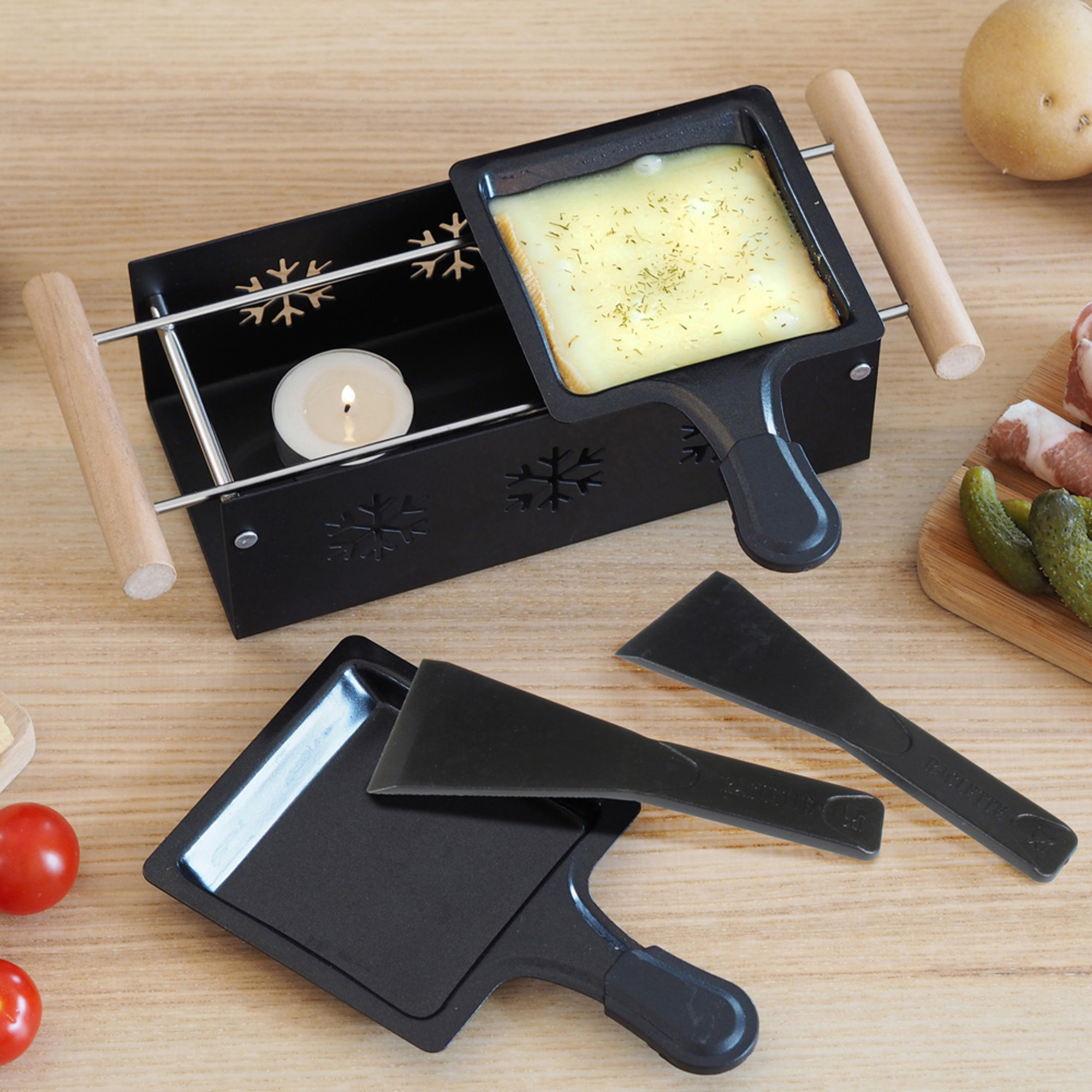 Griglia per raclette nera con formaggio, candela e spatole su legno. Verdure sullo sfondo.