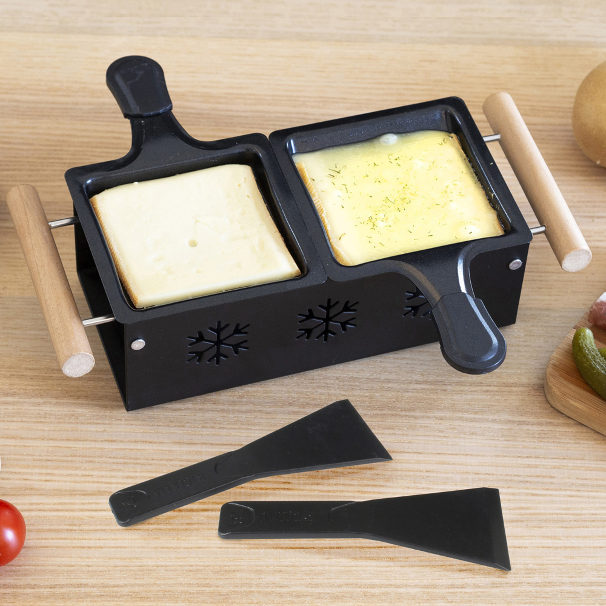 Grill per raclette con formaggio e spatole. Formaggi si sciolgono. Manici in legno e sfondo bianco.