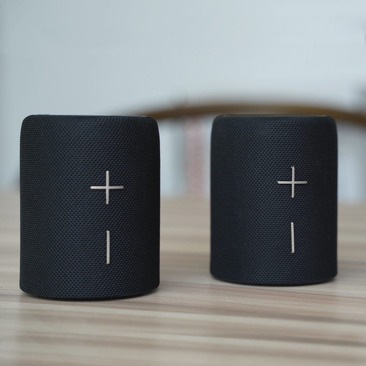 Dwa czarne głośniki ze znakiem plus z przodu, umieszczone na drewnianej powierzchni.