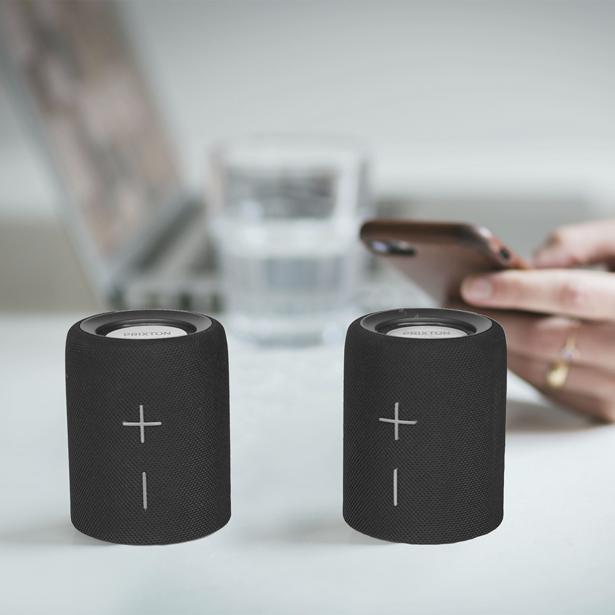 Dwa czarne głośniki ze znakiem plus i linią. Kobieta z telefonem i laptopem w tle.