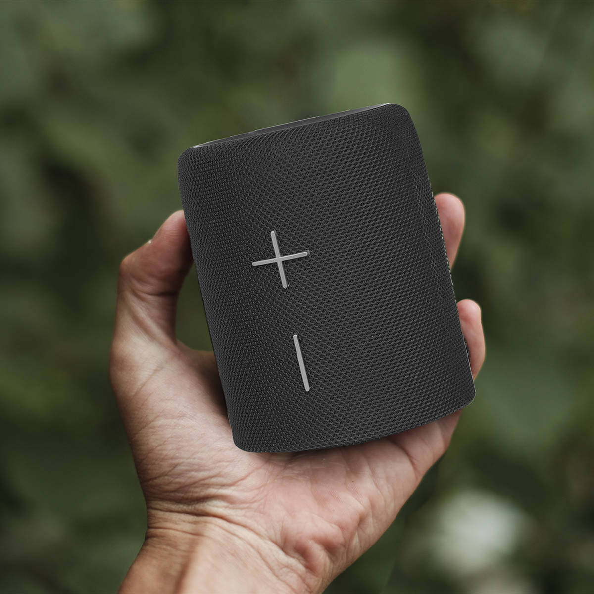 Ręka trzymająca czarny głośnik ze znakami plus i minus. Tło to rozmyte zielone liście.