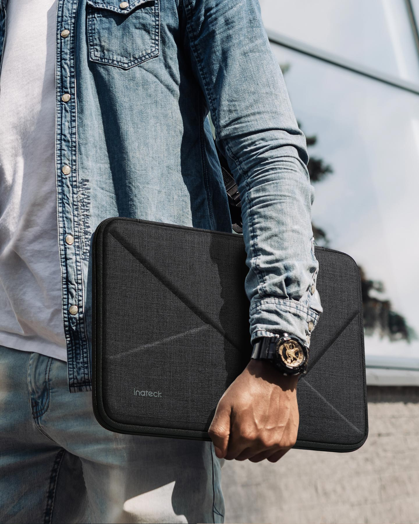 Person hält eine schwarze Laptop-Hülle. Jeansjacke, Uhr mit goldenen Details und Gebäude.