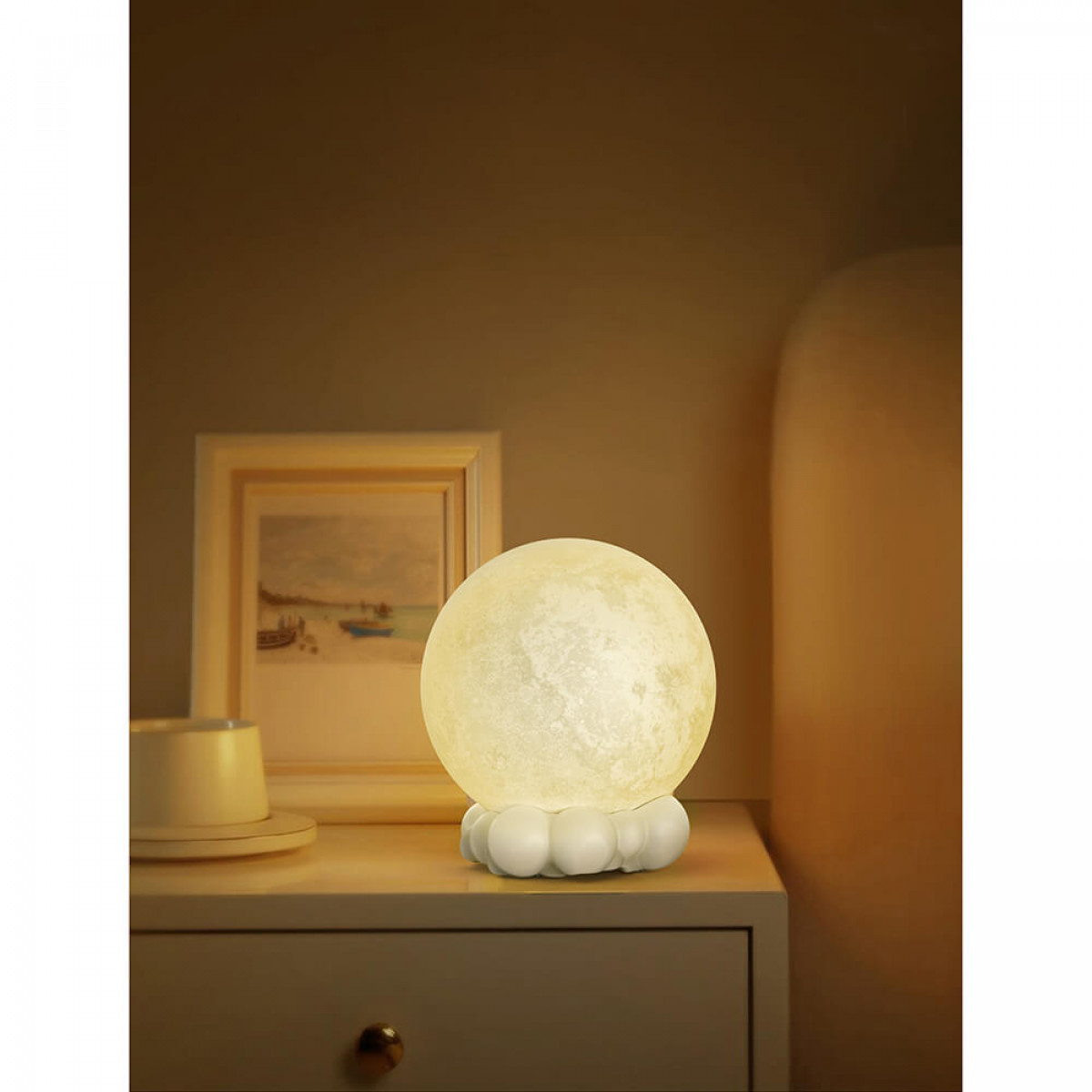Mondförmige Lampe auf einem Nachttisch. Die Lampe leuchtet, mit einem Bild und einer Tasse in der Nähe.