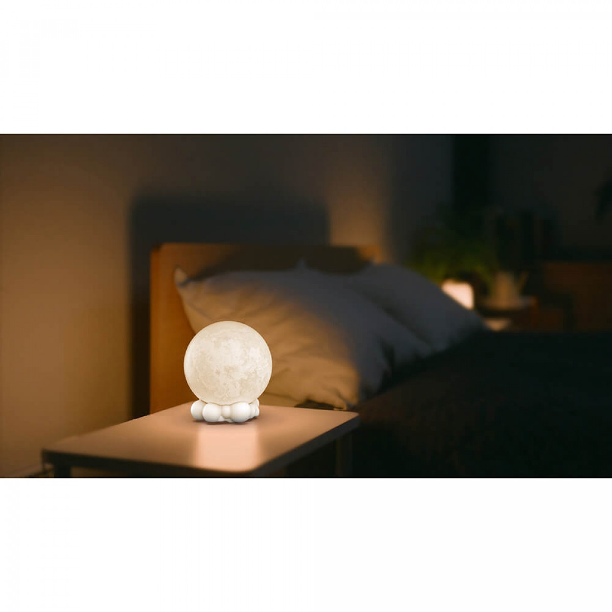 Mondförmige Lampe auf einem Nachttisch. Bett und Kissen im Hintergrund. Dunkle Umgebung.
