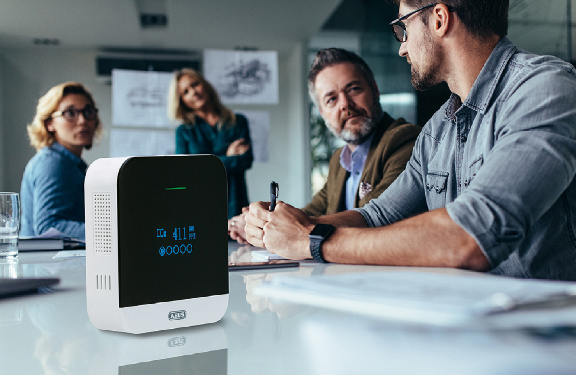 Personen sitzen um einen Tisch, ein weißer Luftqualitätsmonitor steht auf dem Tisch.