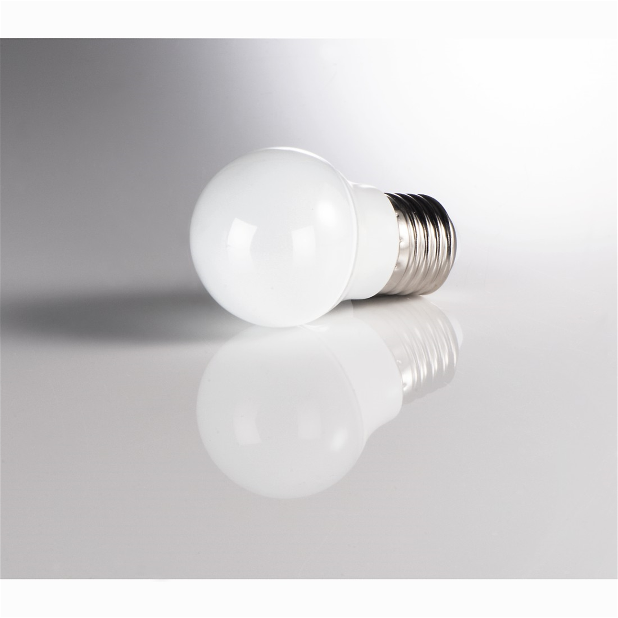 Una lampadina bianca con base argentata, su una superficie bianca riflettente.