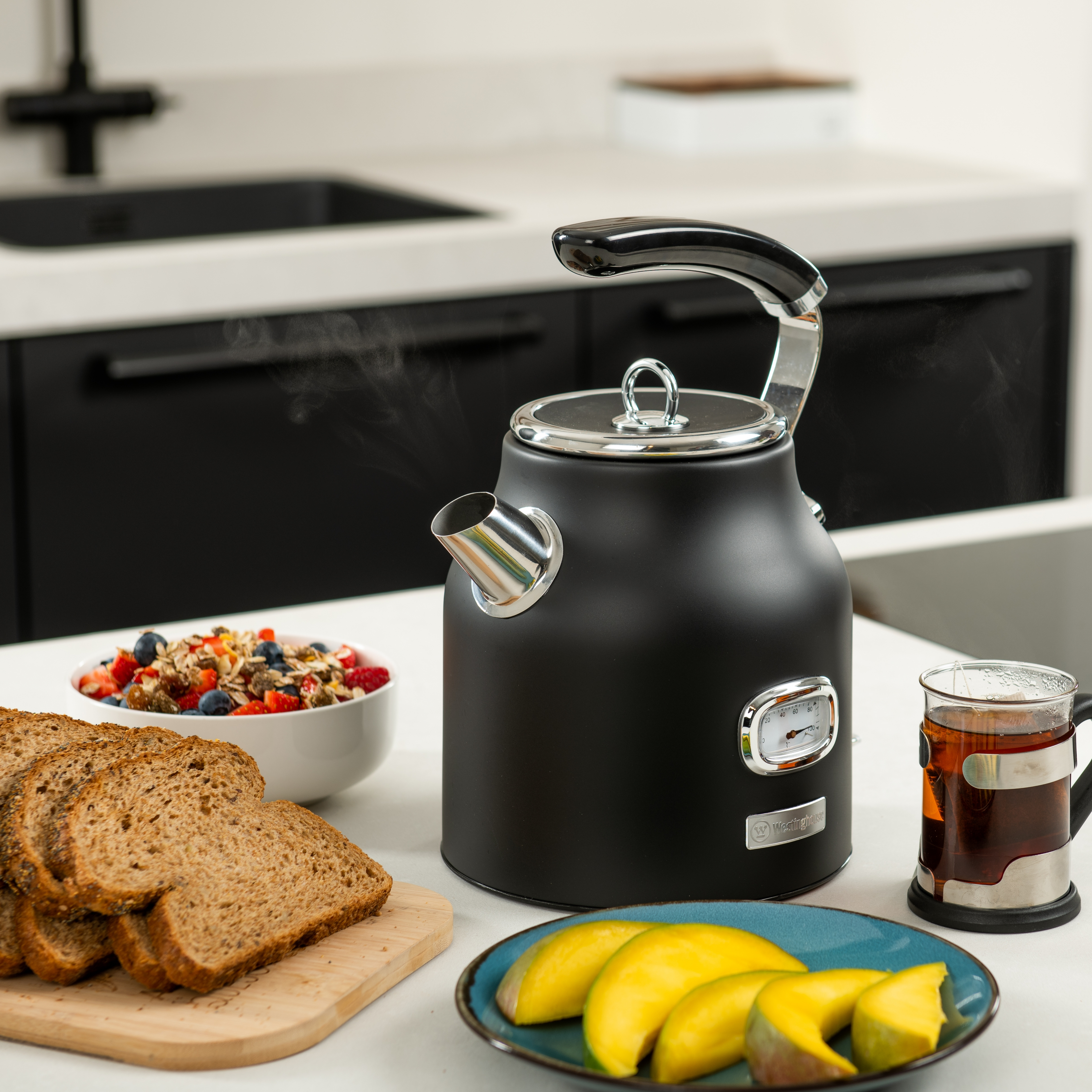 Ein schwarzer Wasserkocher mit Dampf und Essen. Brot, Mangoscheiben und eine Tasse Tee stehen auf dem Tisch.
