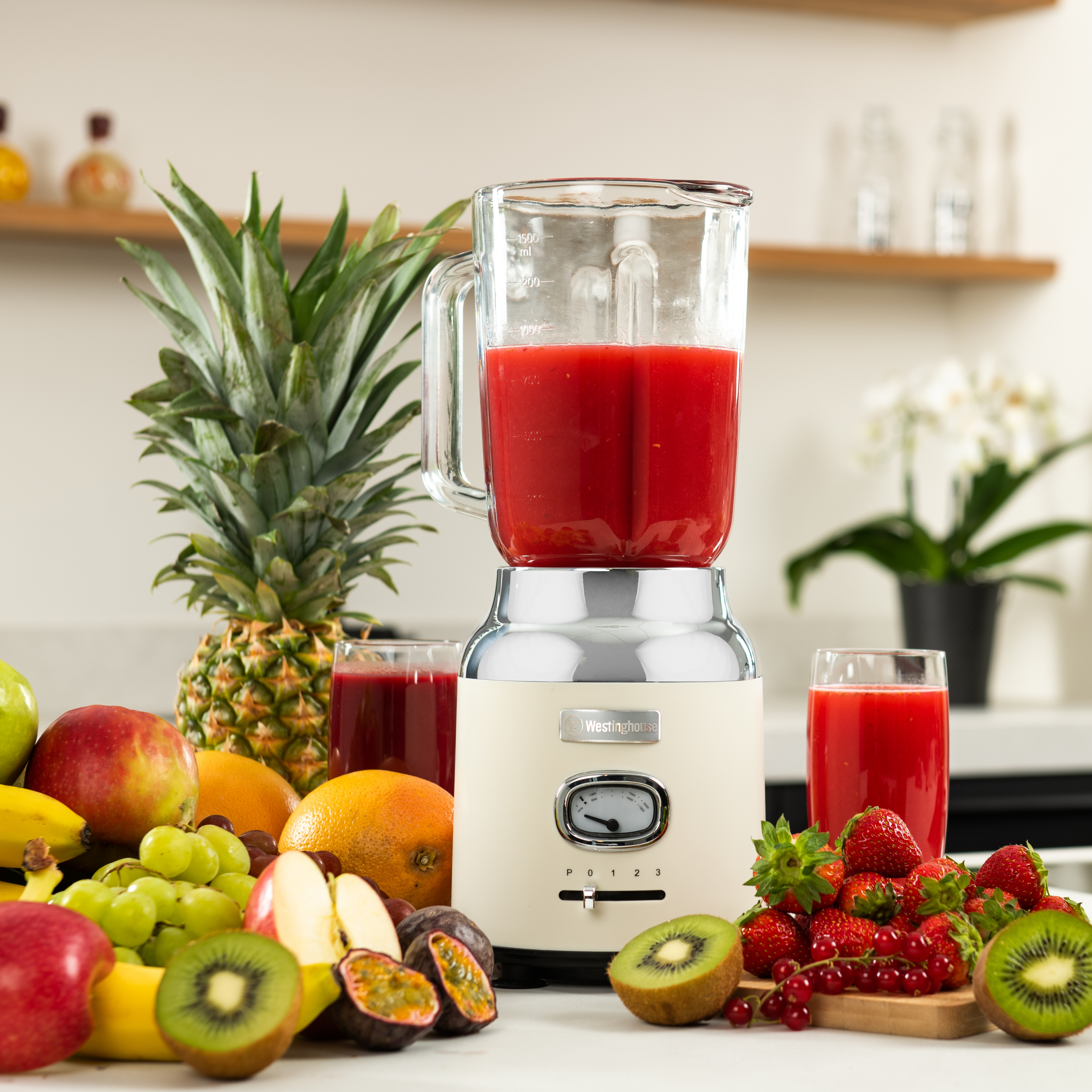 Mixer mit roter Flüssigkeit, Früchten und Gläsern, auf weißem Tisch, Küchenhintergrund.