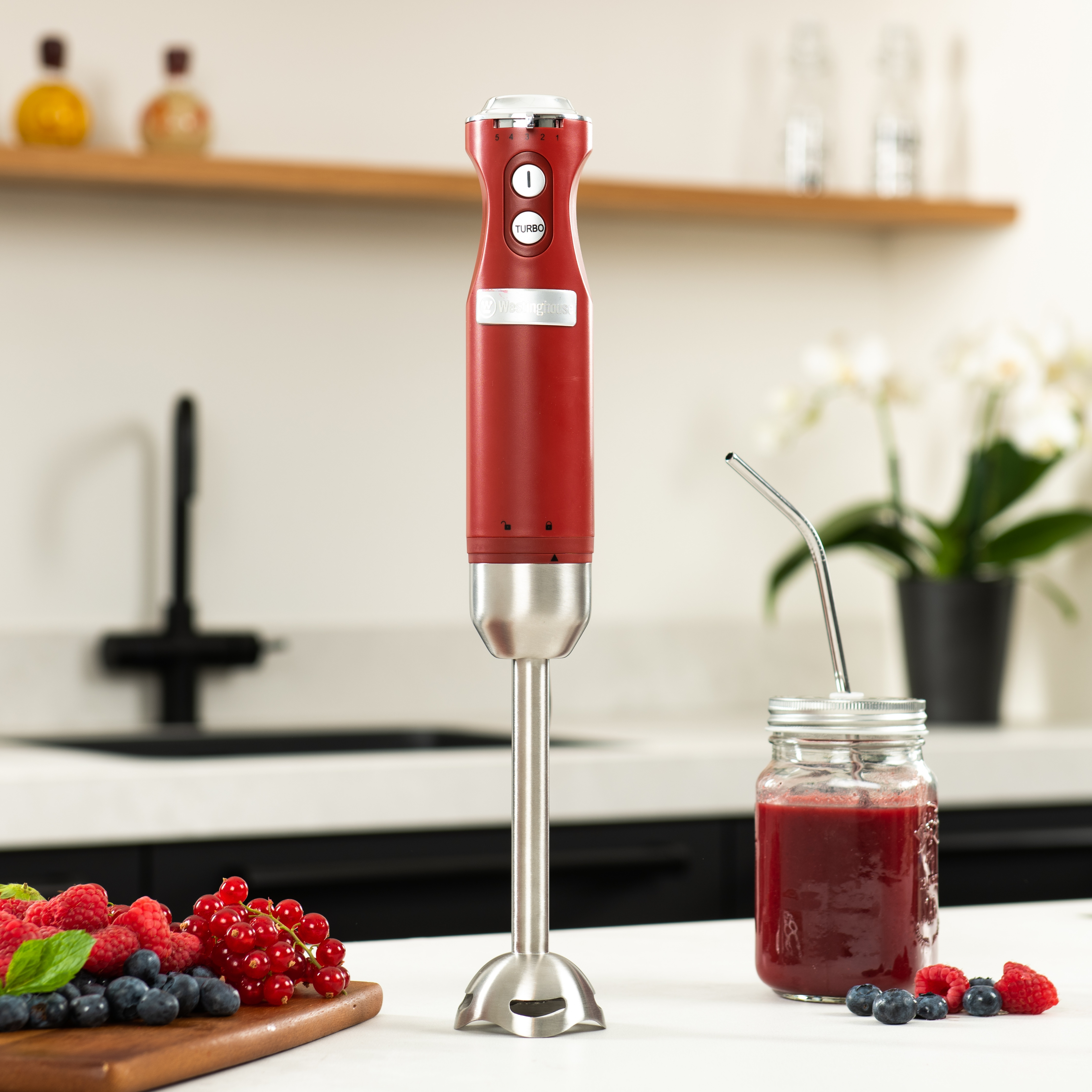 Roter Stabmixer, ein Glas mit Smoothie und Beeren auf einem Holzbrett in einer Küche.
