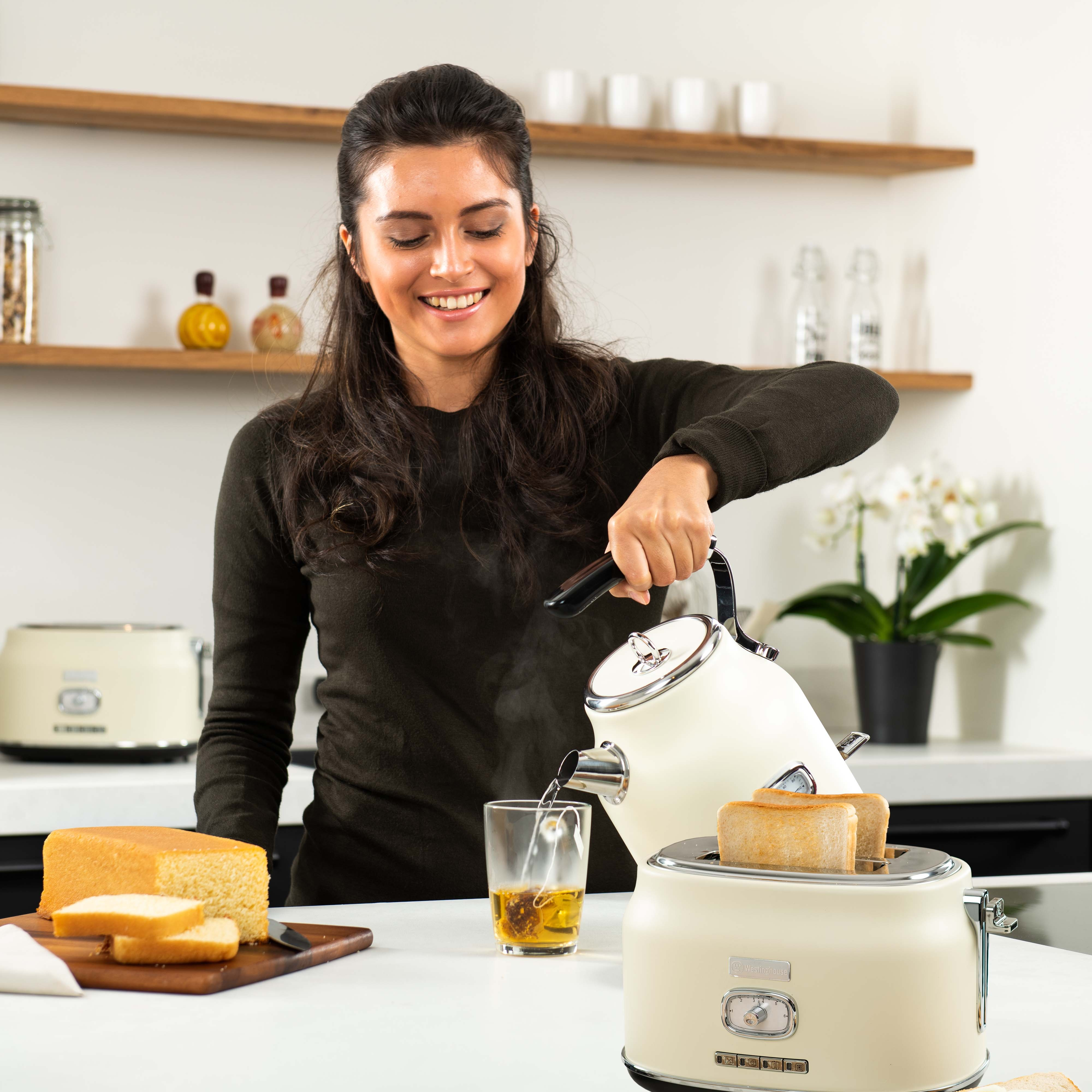 Frau gießt Wasser aus einem weissen Wasserkocher in ein Glas. Toaster und Kuchen.