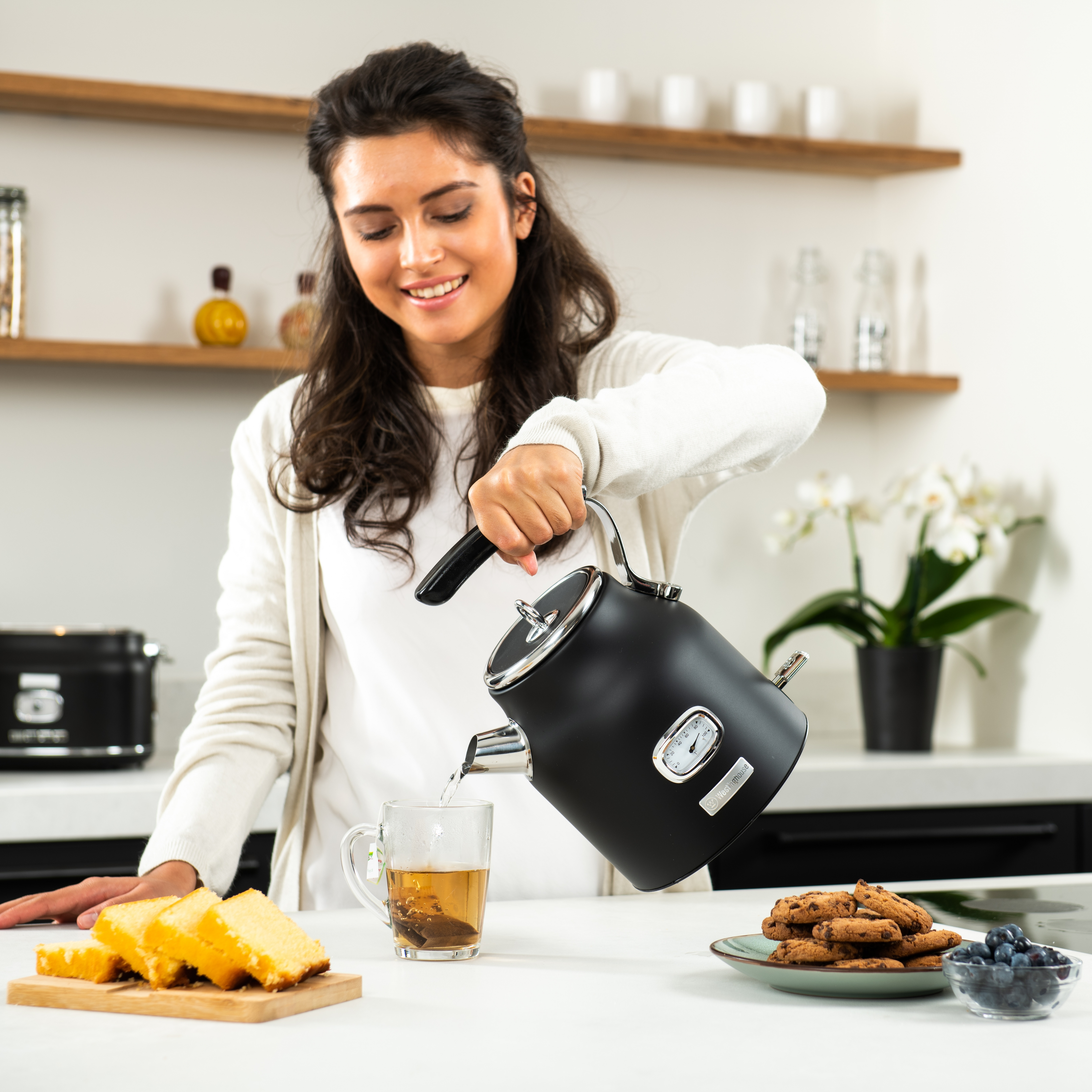 Eine Frau gießt Wasser aus einem schwarzen Wasserkocher in eine Teetasse in einer Küche.