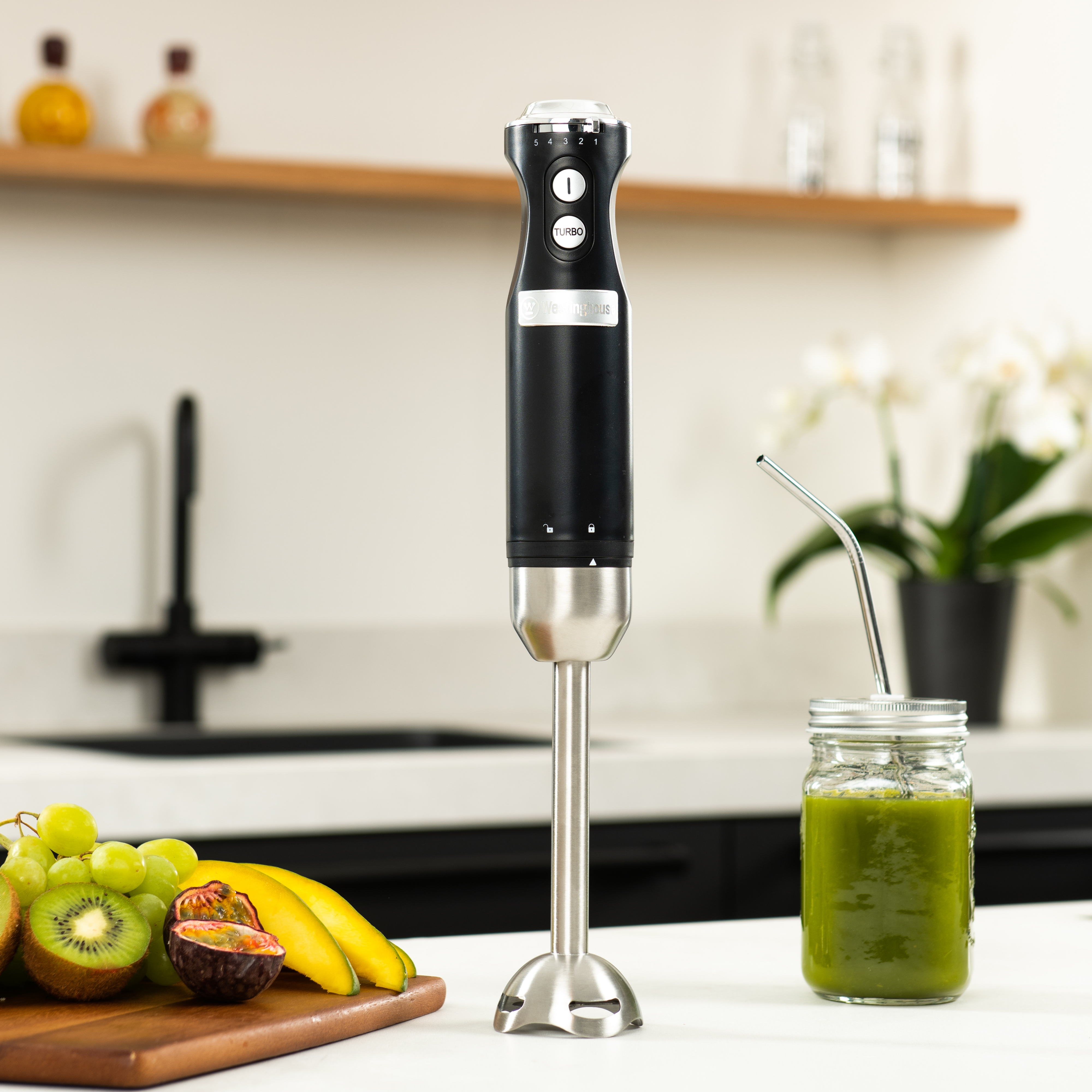 Ein schwarzer Stabmixer mit Edelstahlschaft und ein Glas mit grünem Smoothie. Hintergrund Küche.
