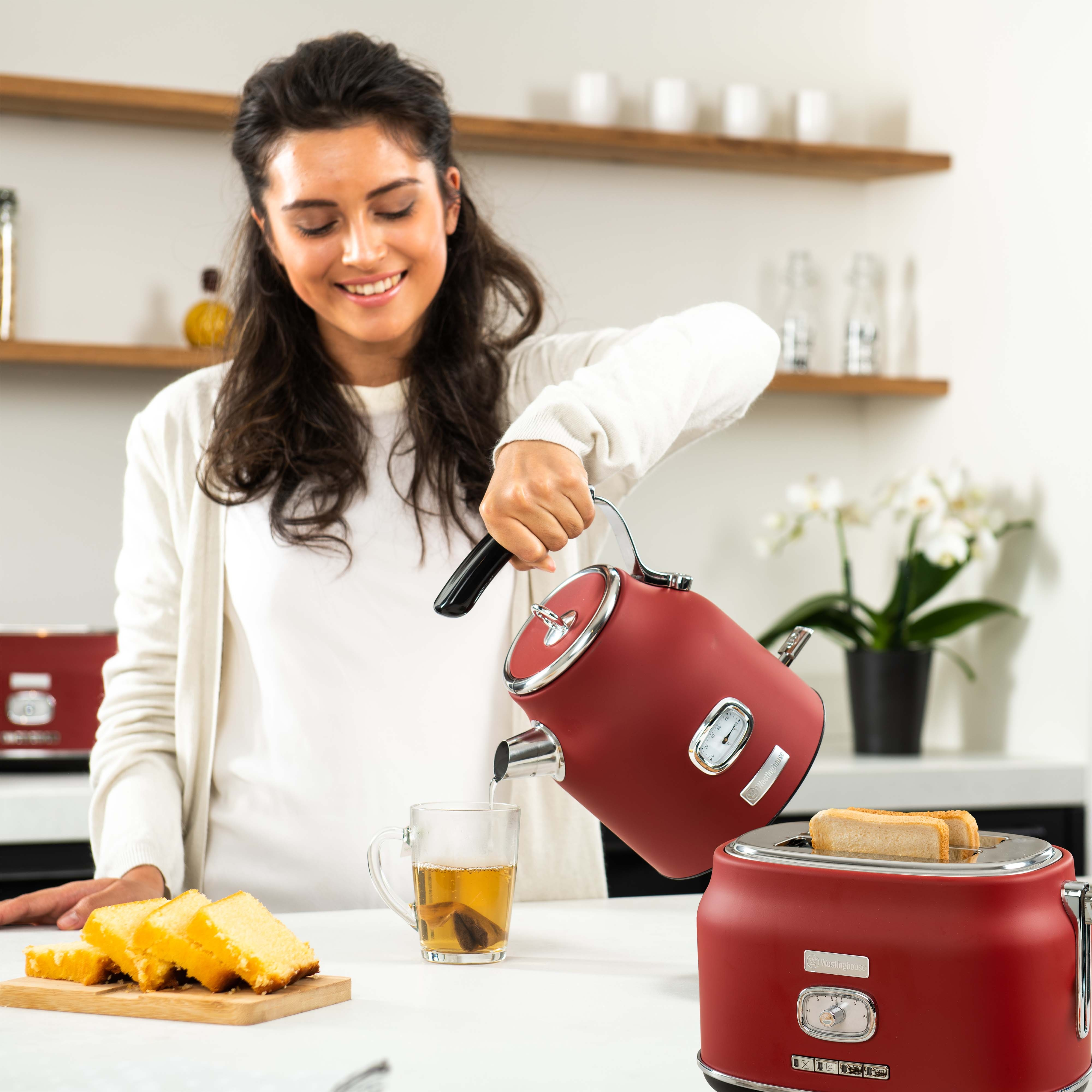 Eine Frau gießt Wasser aus einem roten Wasserkocher in eine Tasse, mit Toaster und Kuchen auf dem Tisch.
