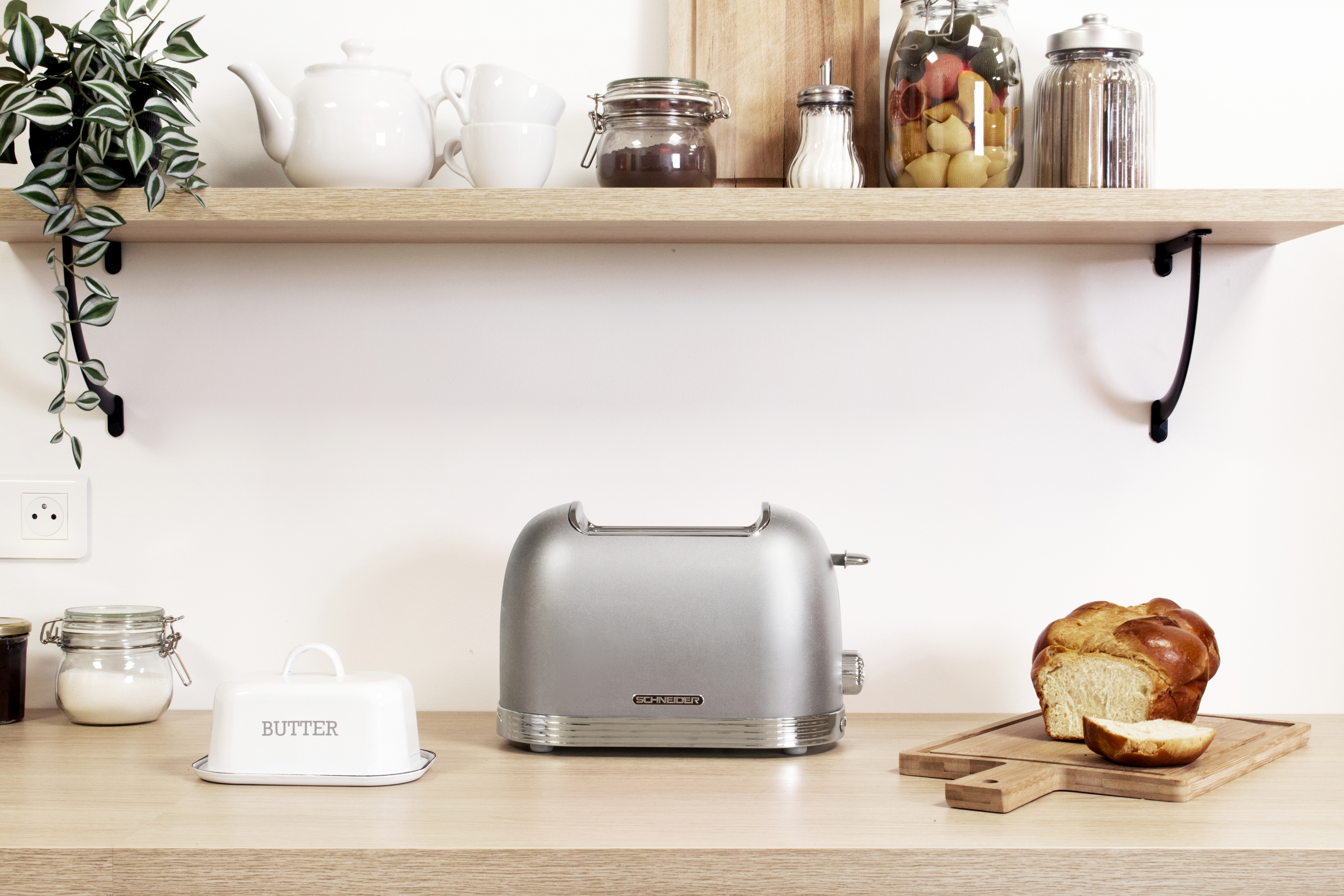 Eine Küchenzeile mit einem silbernen Toaster, einer Butterdose und einem Brotlaib auf einem Holzbrett.
