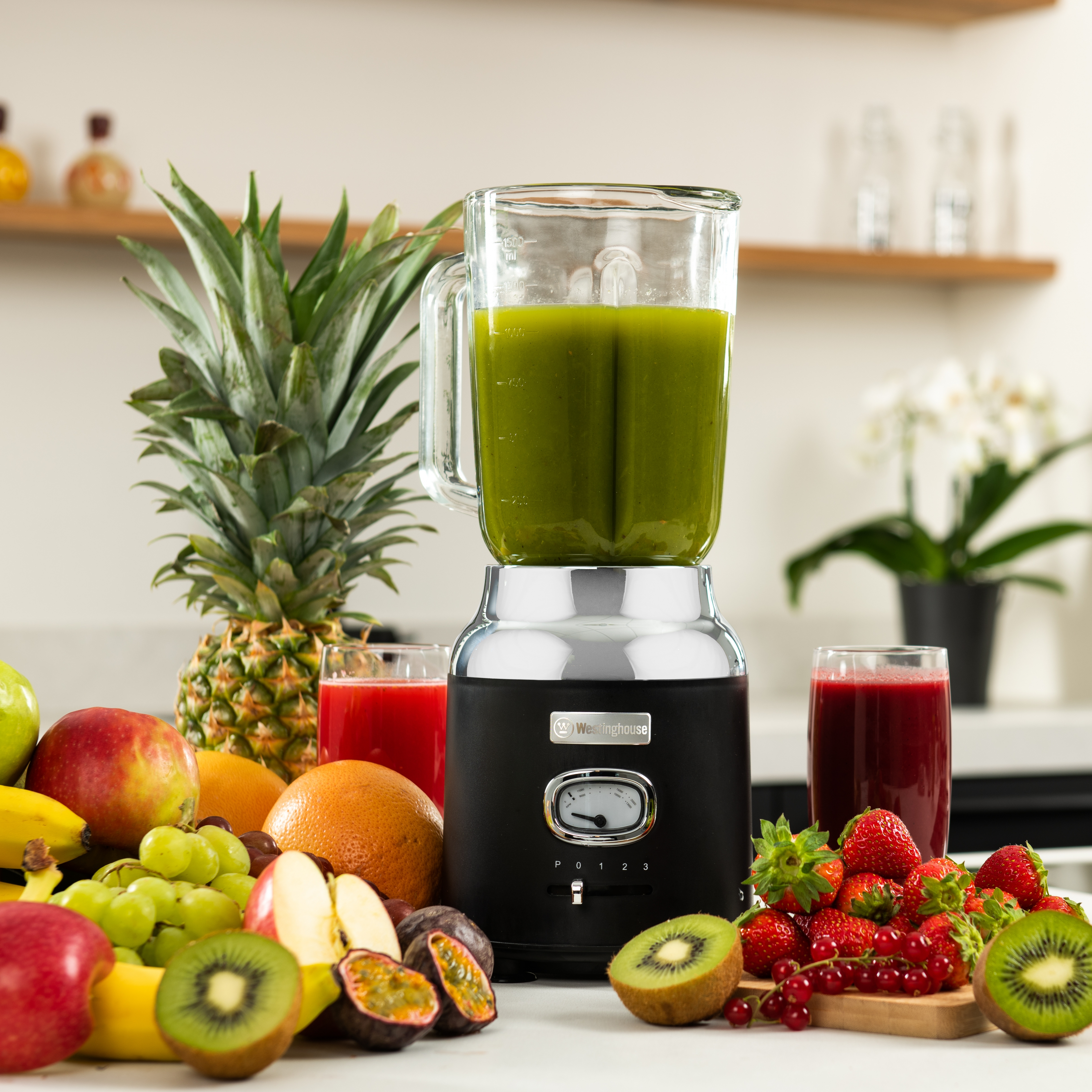 Mixer mit grünem Smoothie, Früchte: Ananas, Äpfel, Kiwi, Erdbeeren. Weiße Oberfläche, verschwommener Hintergrund.