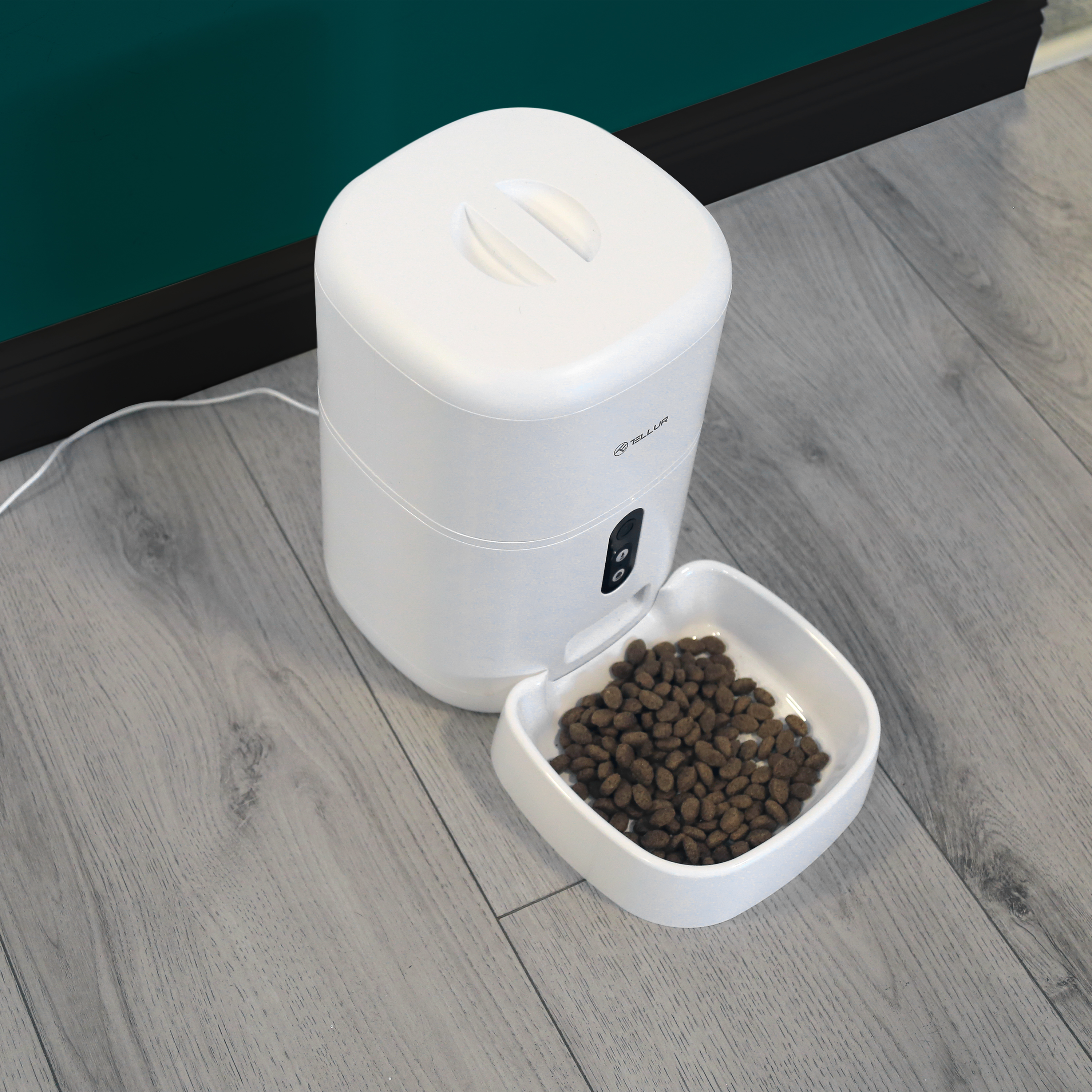 Weißer Tierfutterautomat mit einer Futterschüssel auf dem Boden, neben einer grünen Wand.