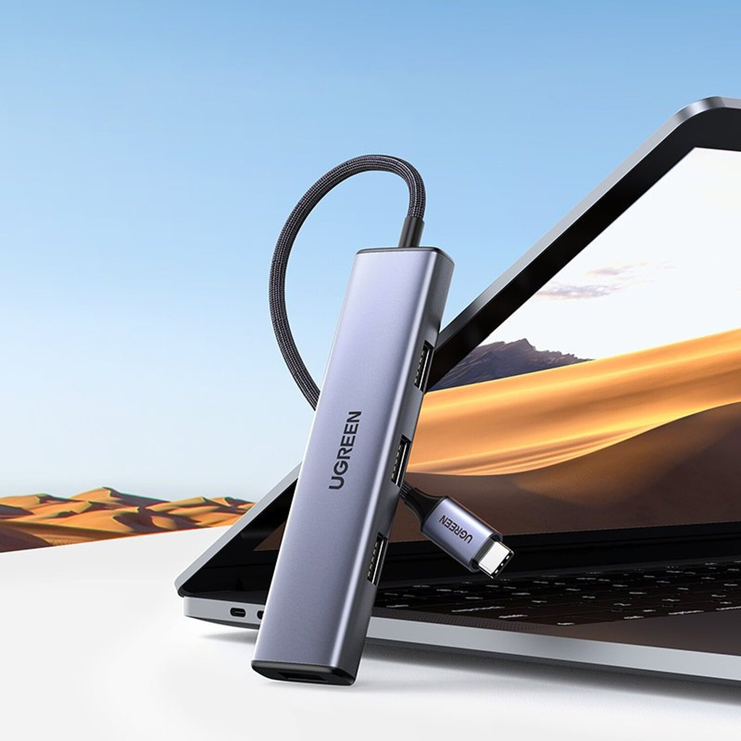 Szary hub USB-C UGREEN jest podłączony do laptopa. Tło przedstawia scenerię pustynną.