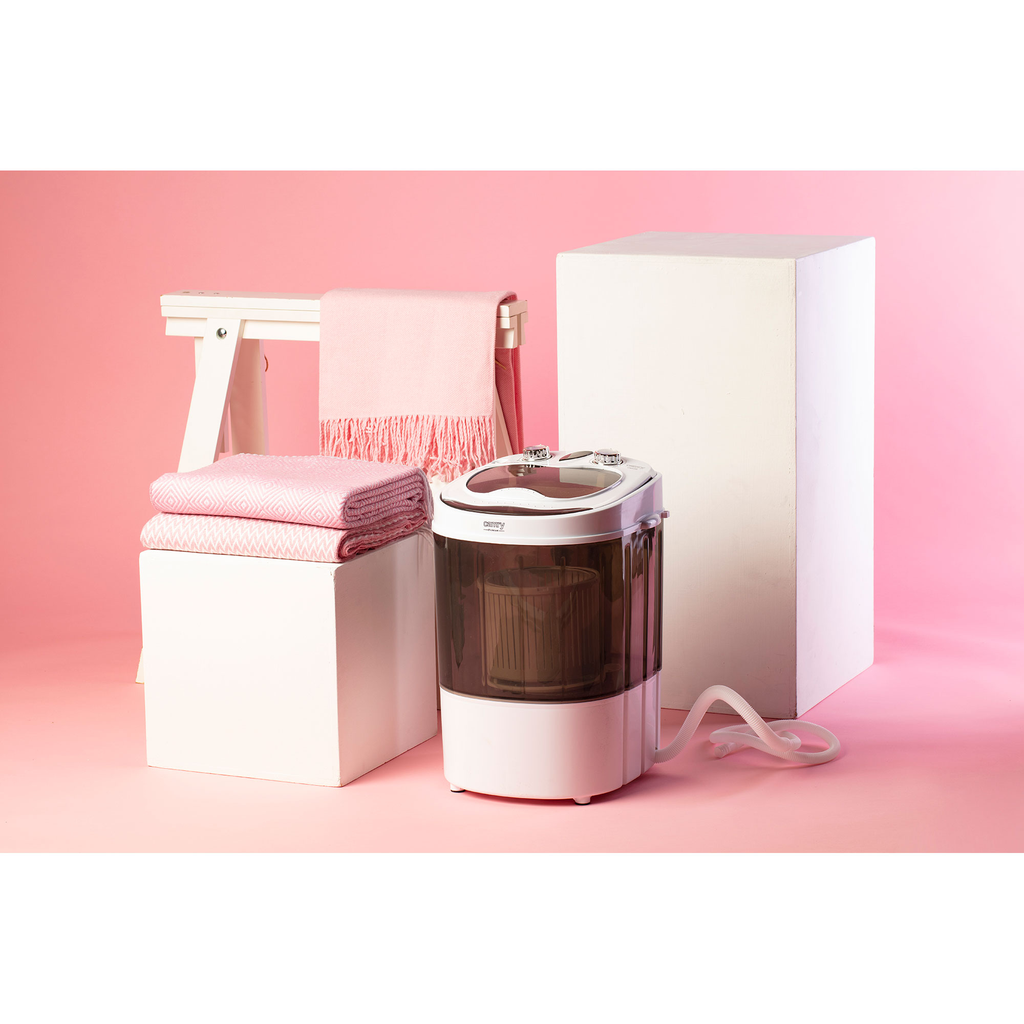 Mała biała pralka stoi obok różowych koców na białym postumencie.