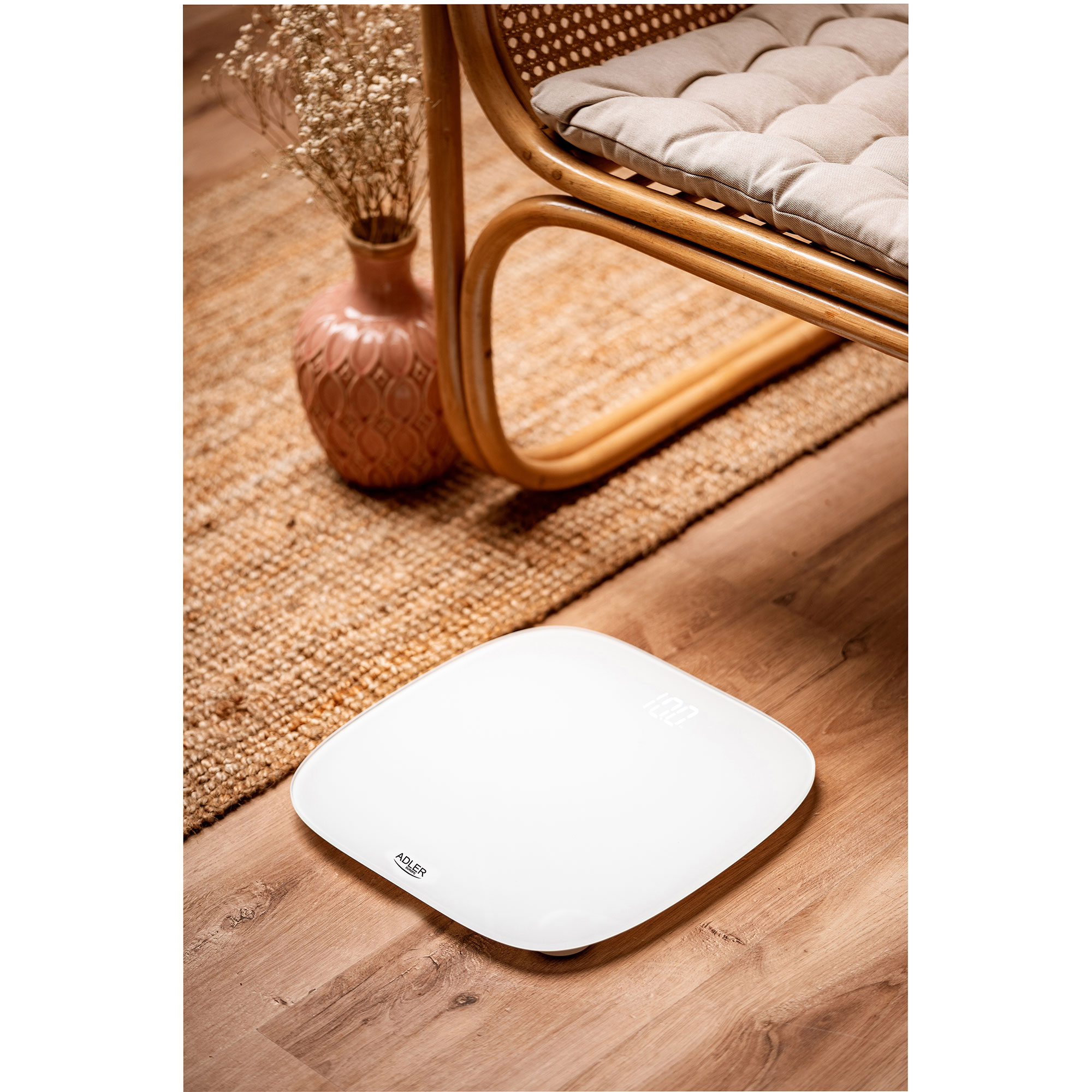 Biała waga na drewnianej podłodze, z krzesłem i wazonem w tle. Waga pokazuje 100.