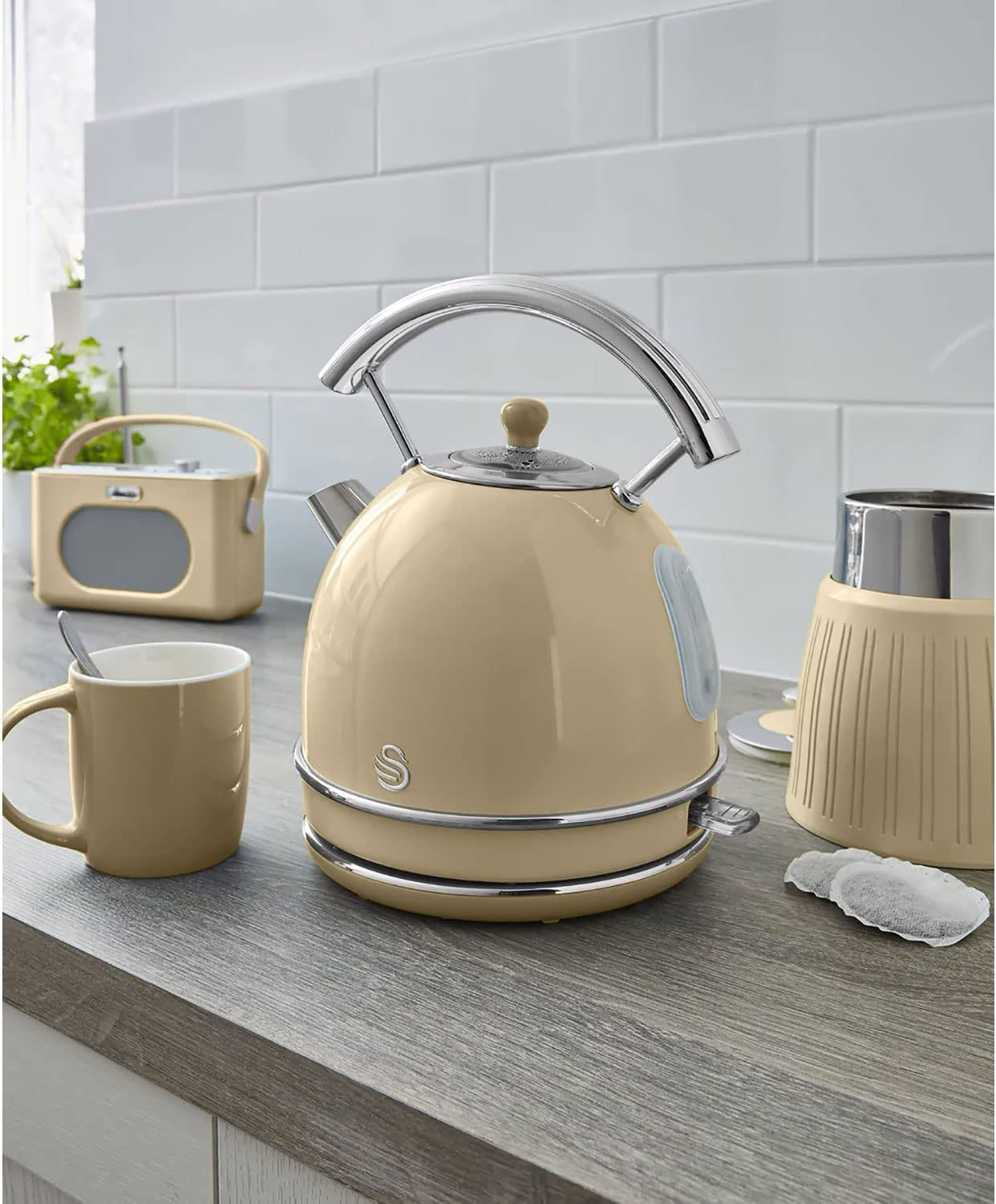 Beige Wasserkocher mit silbernem Griff, Tasse, Radio und Mixer auf Holz-Arbeitsplatte.
